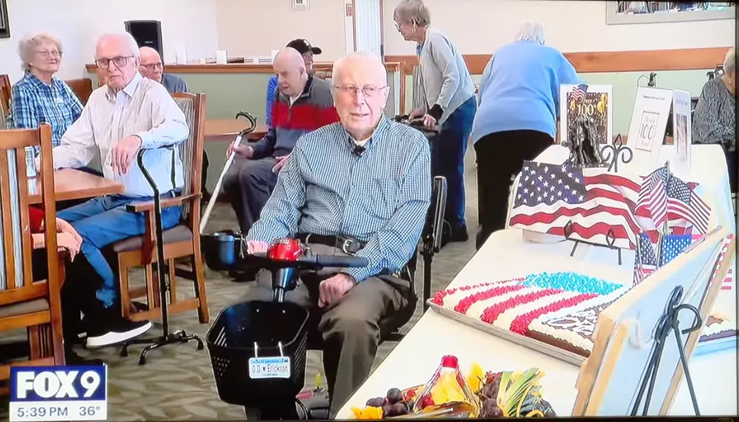 Elderly man sitting in a wheelchair in a room with other seniors, tables, and decorations, including American flags and patriotic items.