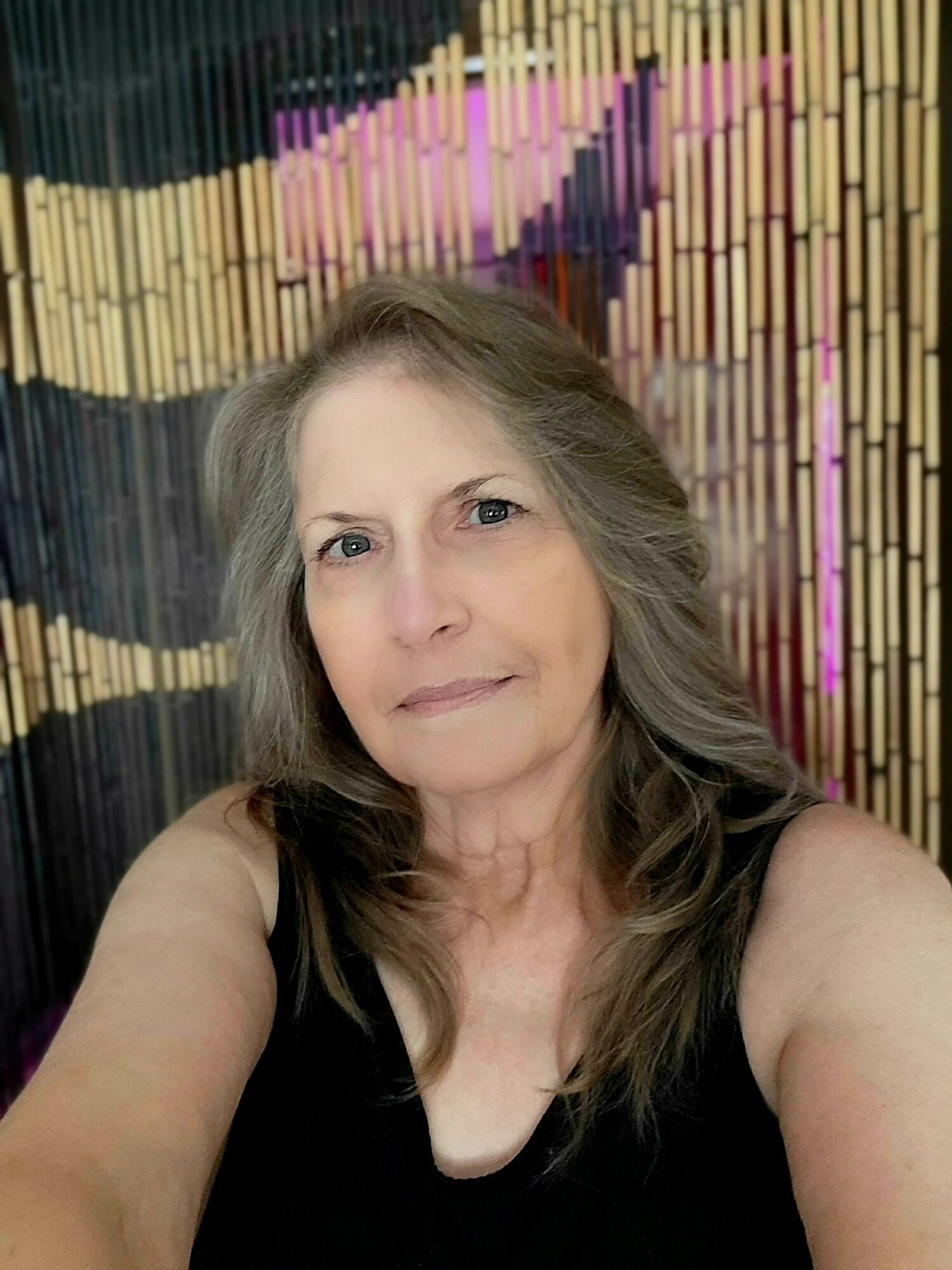 Woman with long gray hair taking a selfie in front of a colorful beaded curtain, wearing a black top.