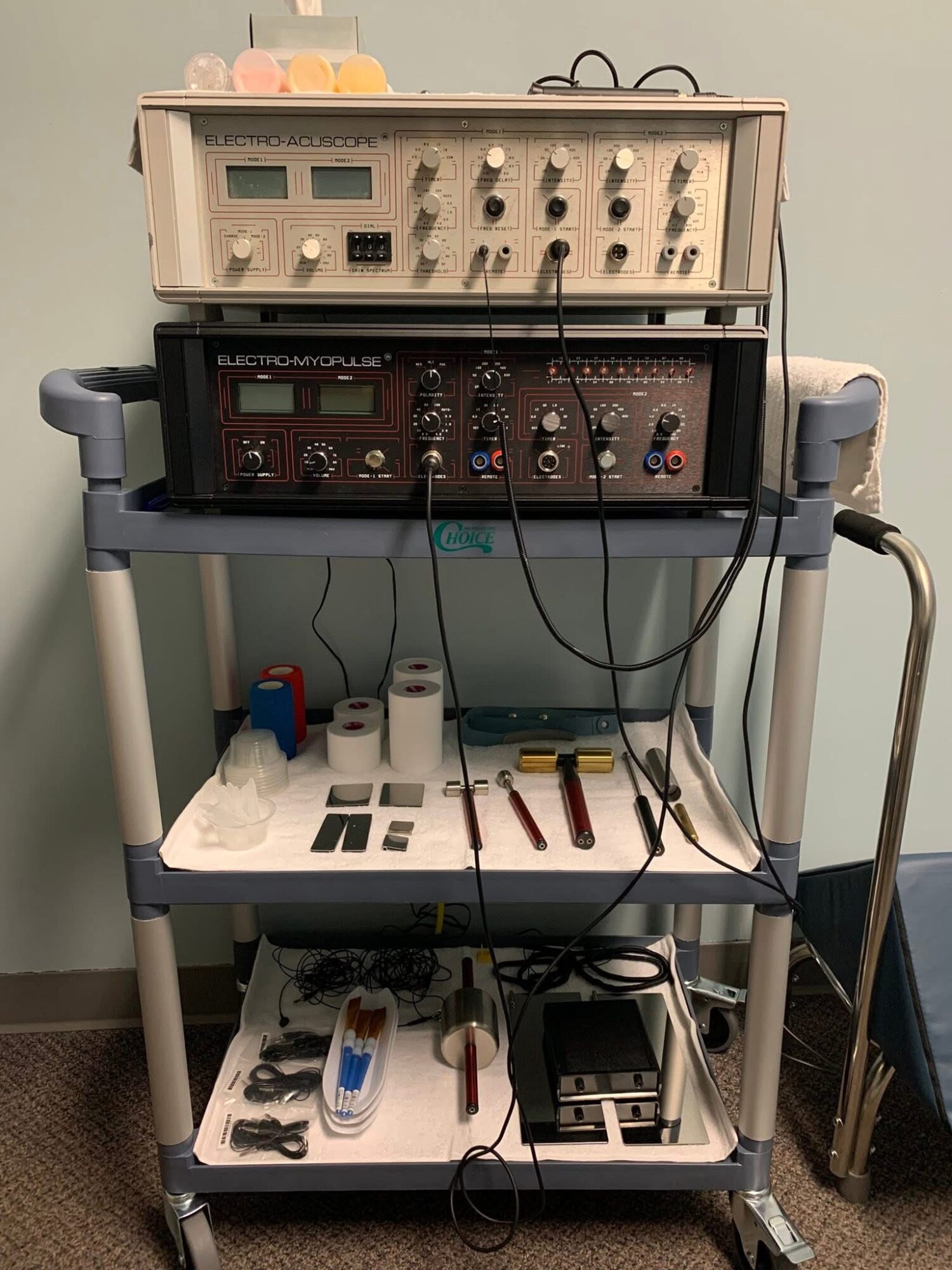 Medical or laboratory equipment on a multi-tiered cart, including electronic devices, tools, and cables, against a plain wall.