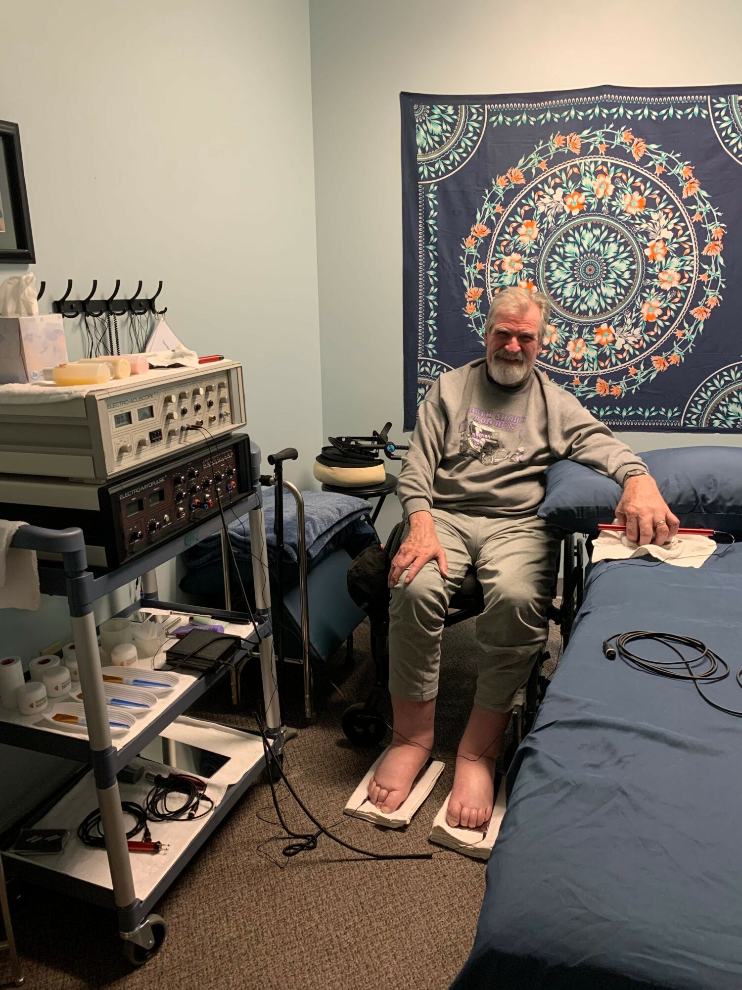 Person sitting on bed holding a book, with electronic equipment and a tapestry on the wall behind.