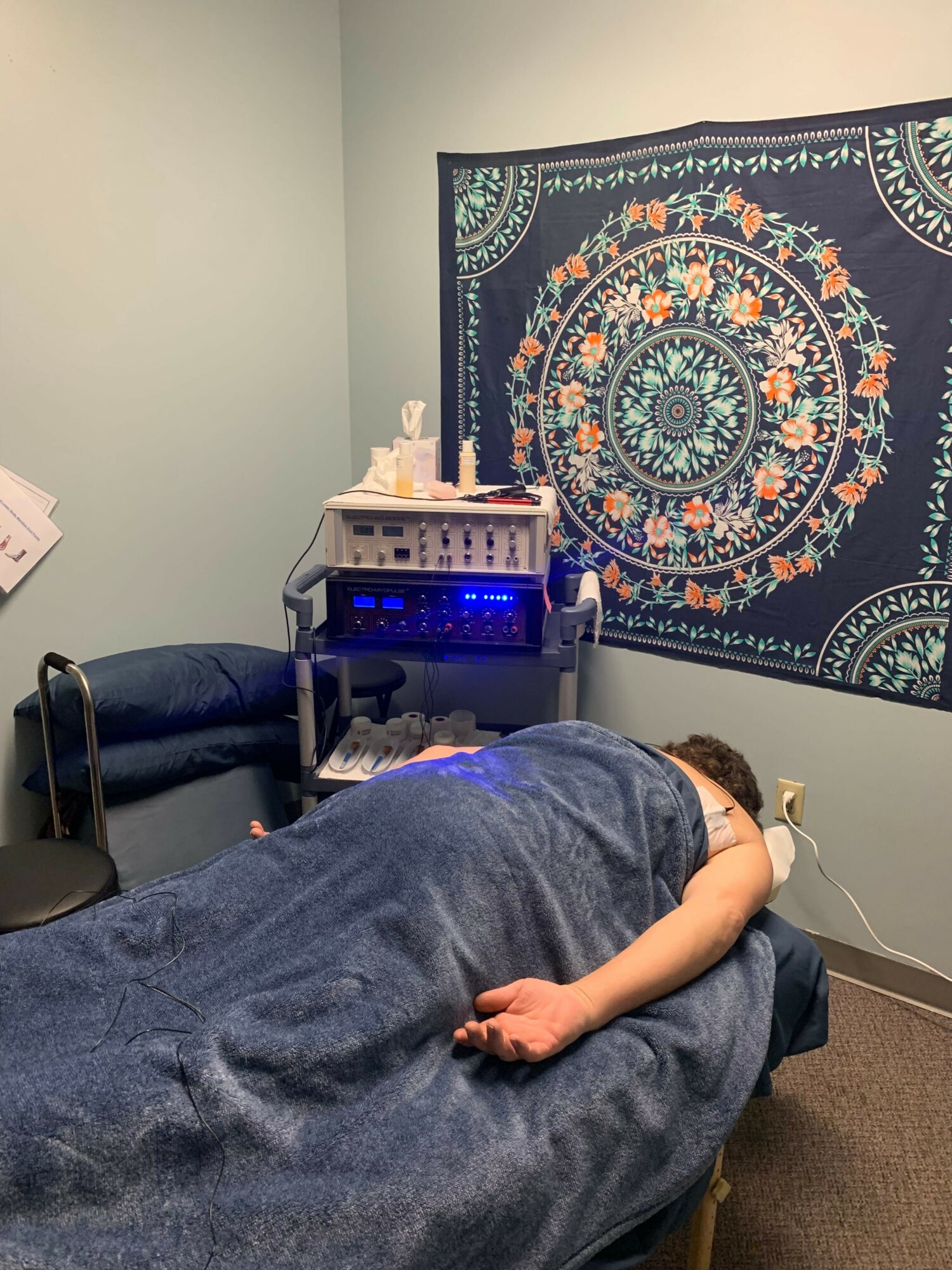 Person lying on a bed with a blue blanket, facing a wall with a colorful mandala tapestry, in a room with electronic equipment.