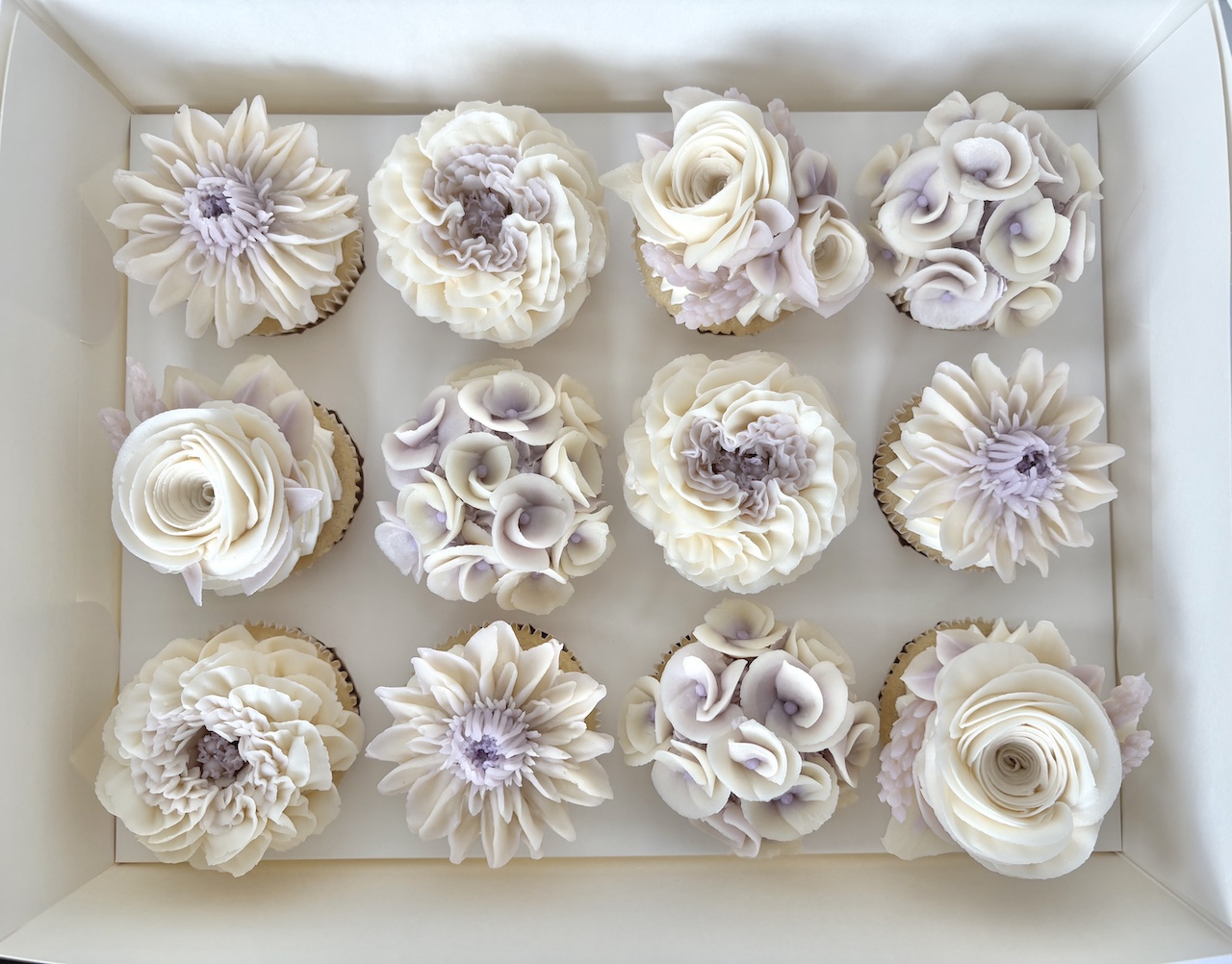 Box of twelve cupcakes decorated with white and purple floral frosting designs, arranged in three rows of four.