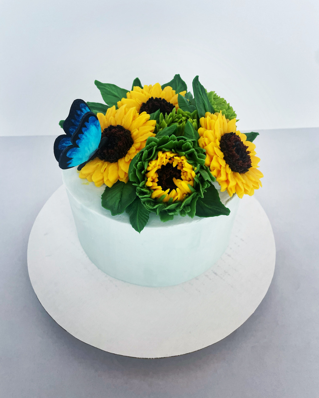 Round white cake with sunflower and green leaf decorations, topped with a blue butterfly, on a white surface.