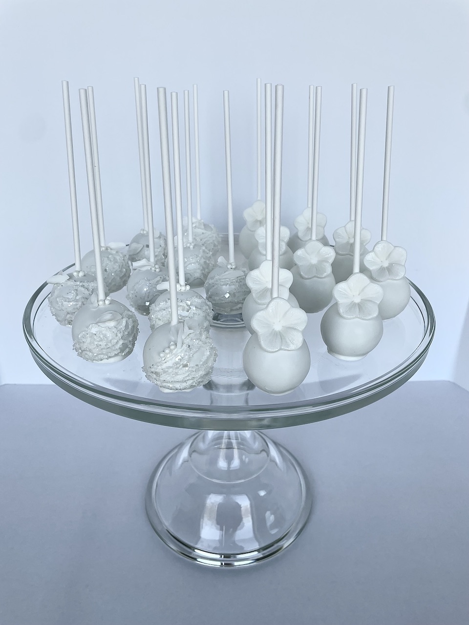 Clear glass cake stand with white and silver cake pops, some decorated with flowers, on a light background.