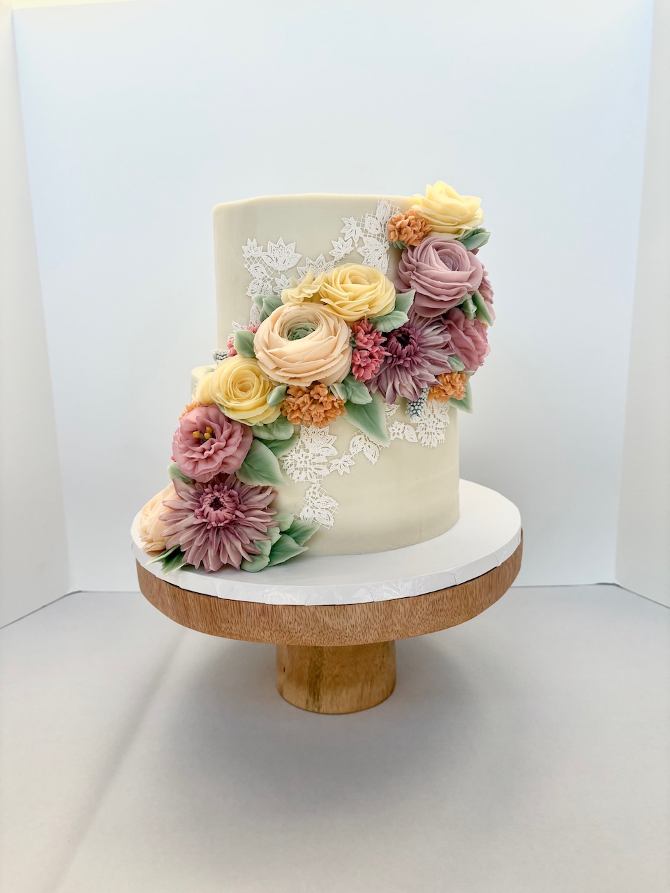 Elegant cake decorated with pastel-colored flowers and lace, on a wooden stand against a plain background.