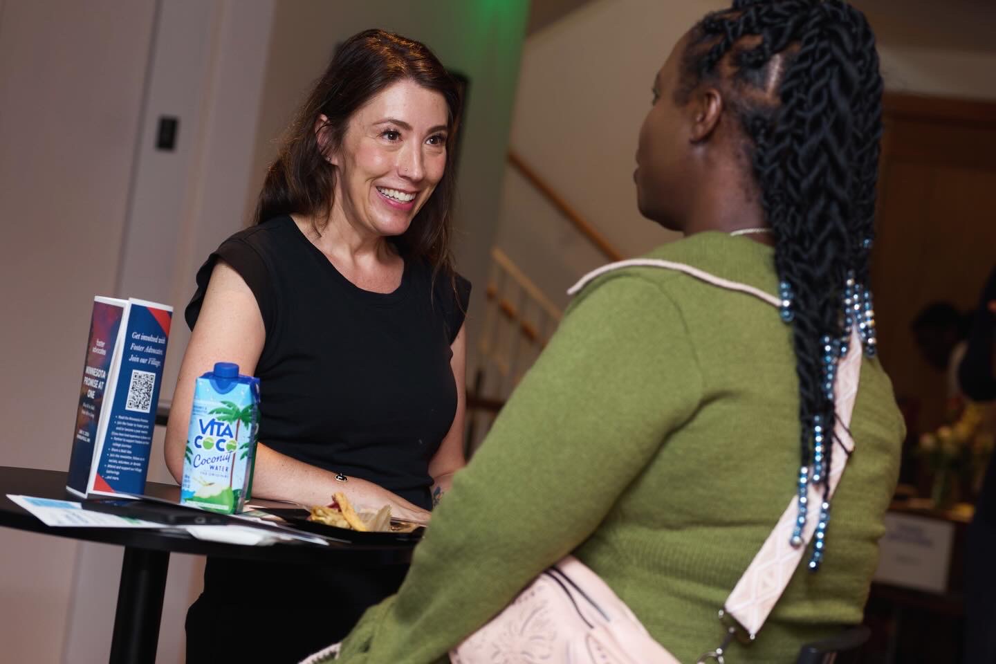 Two women engaged in conversation, smiling, indoors with a table and items including a water bottle and a box.
