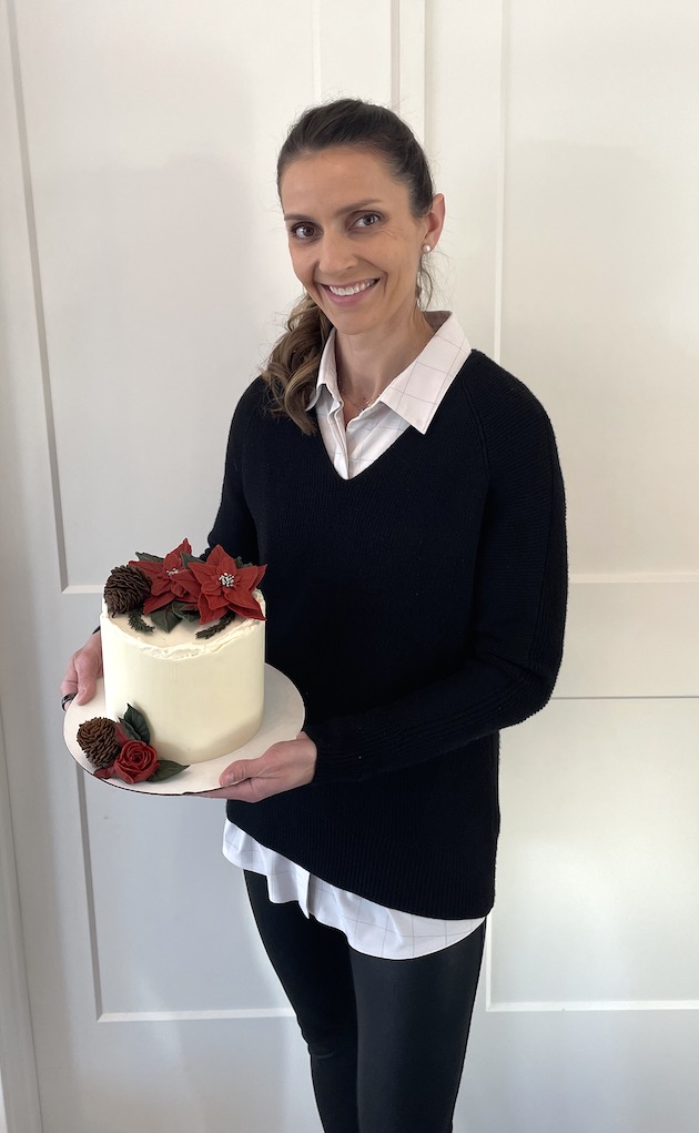 Woman holding a decorated cake with red flowers and berries, smiling, standing against a white background.