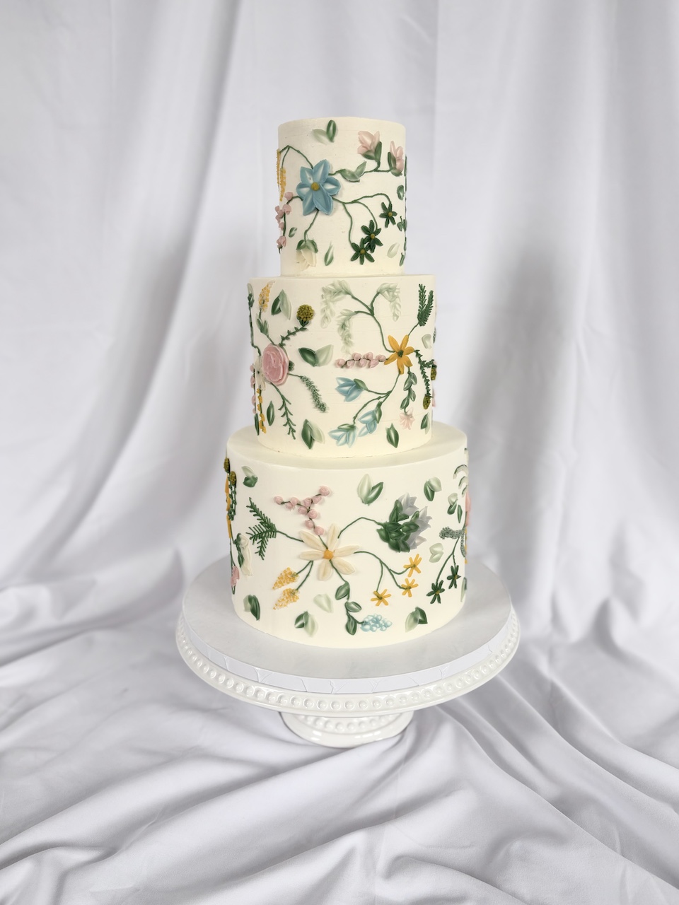 Three-tiered cake decorated with colorful floral and botanical patterns, placed on a white stand against a white fabric backdrop.