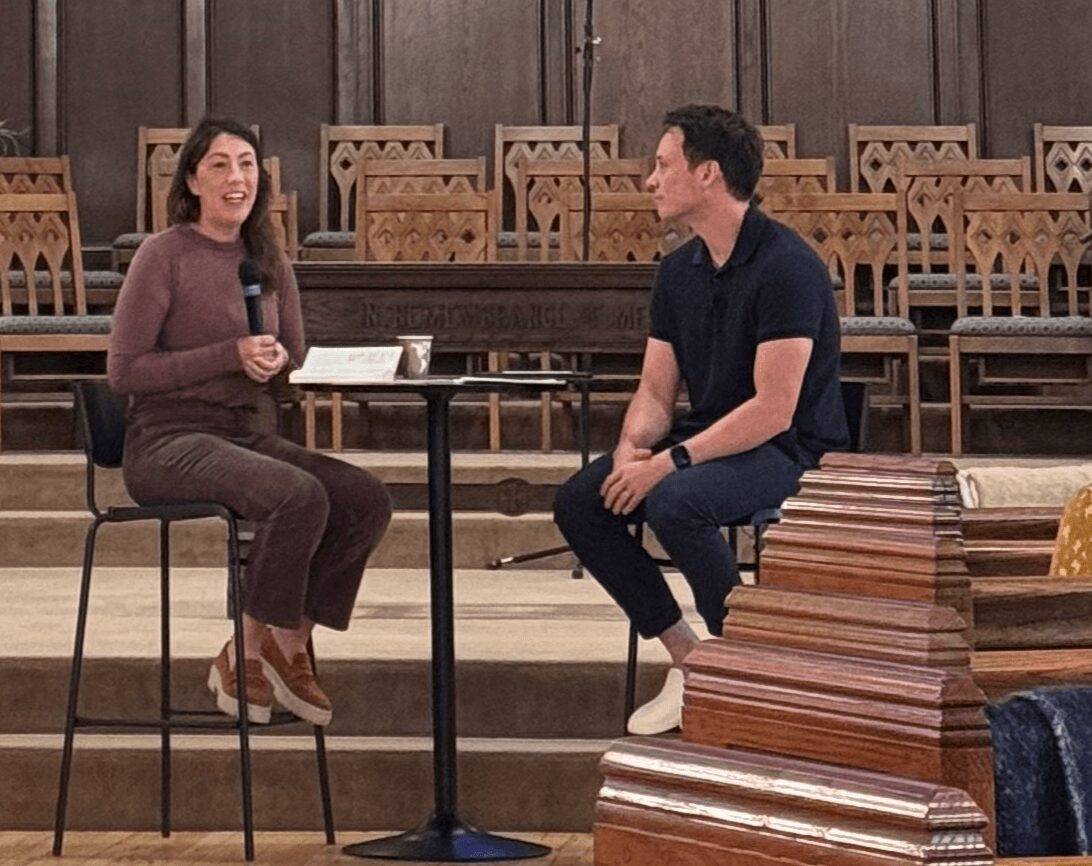 Two people sit on stage, engaging in conversation, with empty chairs and wooden paneling in the background.