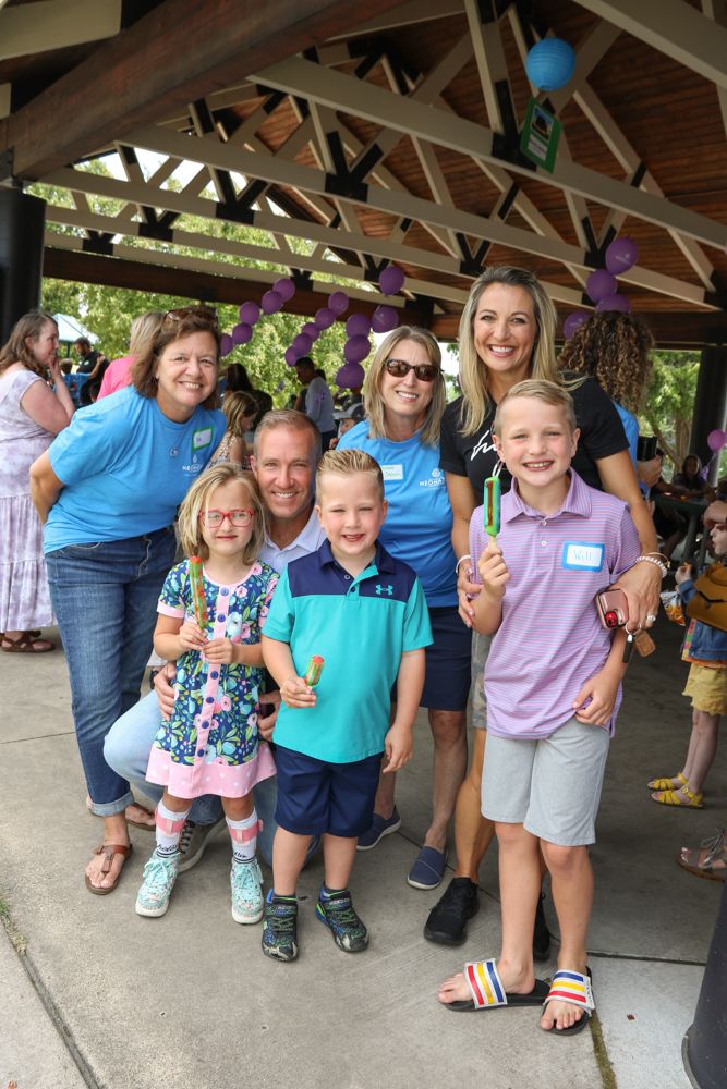 Group of children and adults smiling outdoors under a decorated pavilion with purple balloons, some holding ice cream cones.
