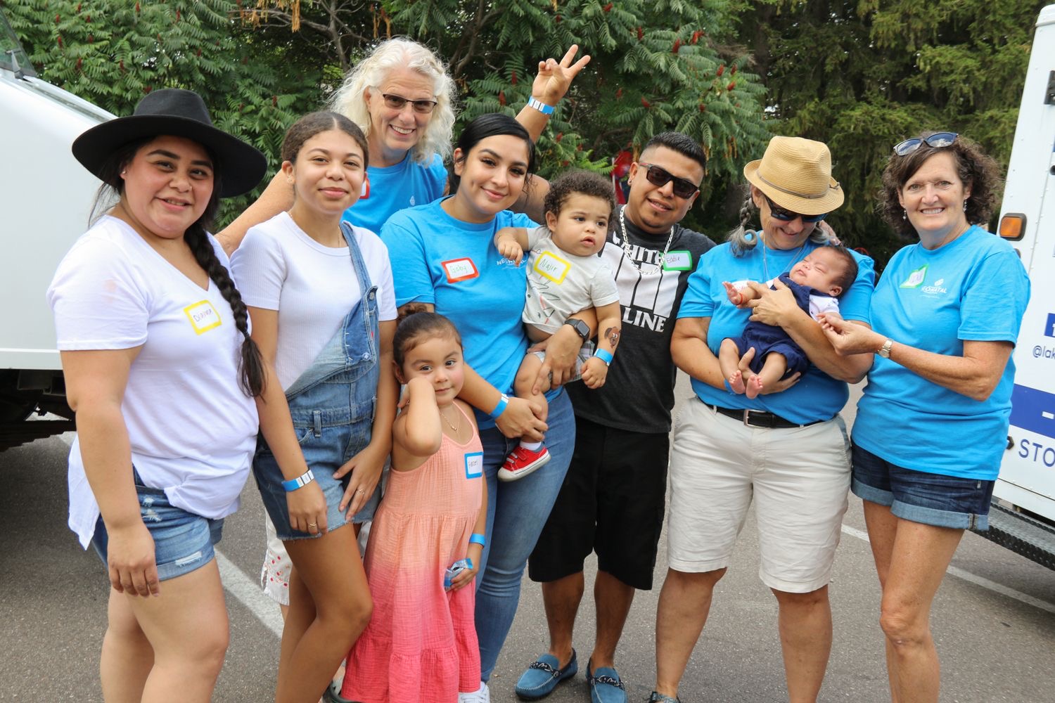 Group of nine people, including children and adults, standing outdoors with trees and a vehicle in the background.