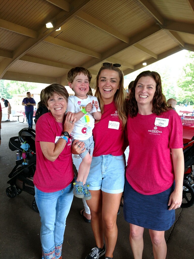 Four people, three women and a child, smiling and standing together under a pavilion, wearing pink shirts, with trees in the background.