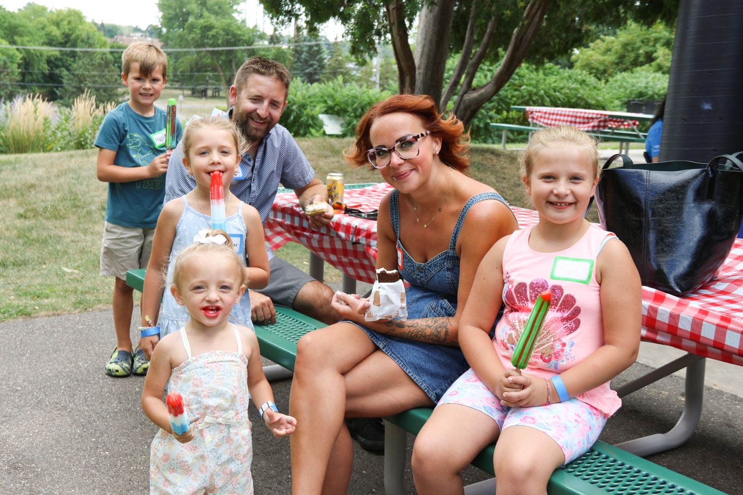Group of children and adults at outdoor picnic table, smiling, with trees and a pond in background.