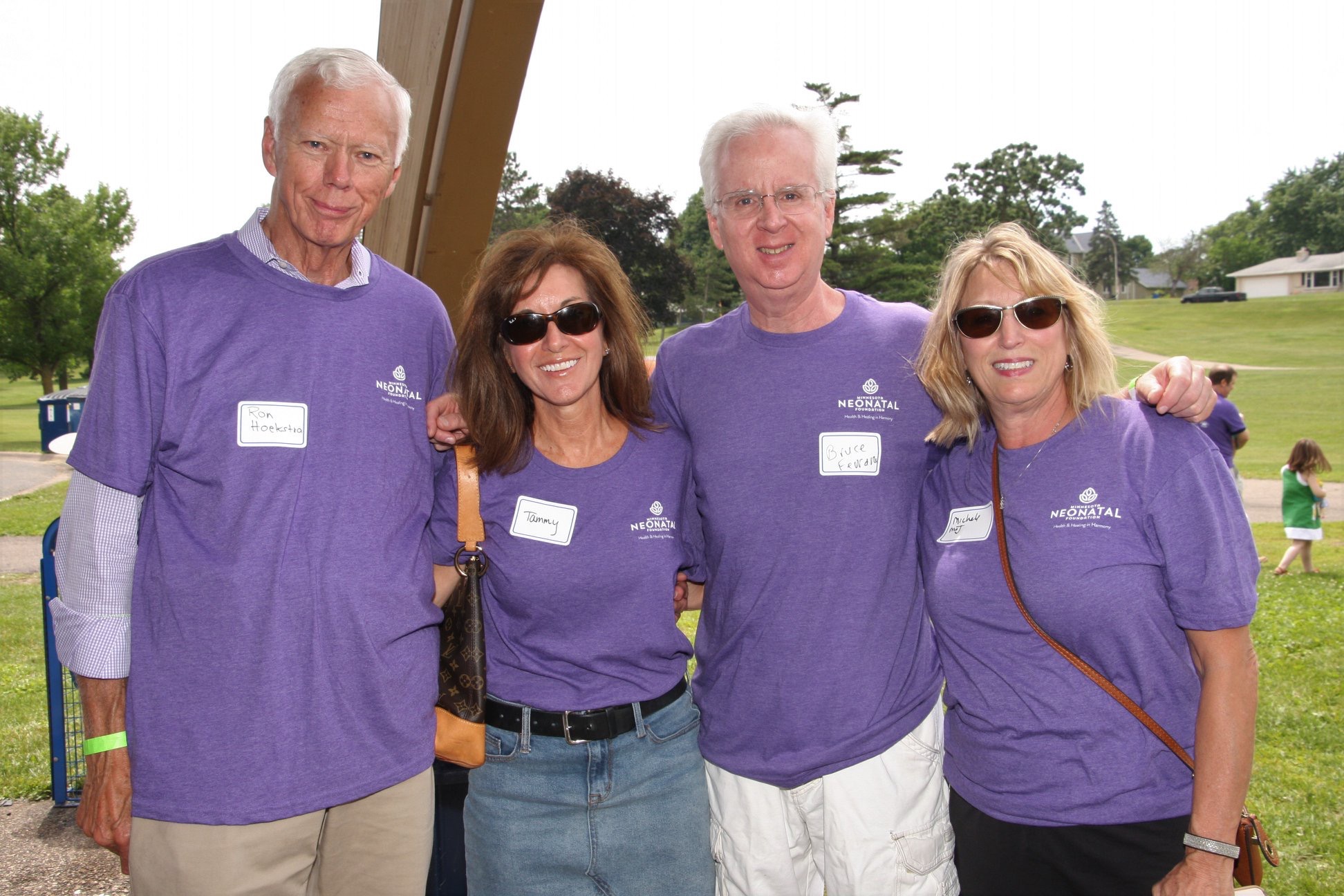 Four people wearing purple shirts standing outdoors, smiling, with trees and a grassy area in the background.