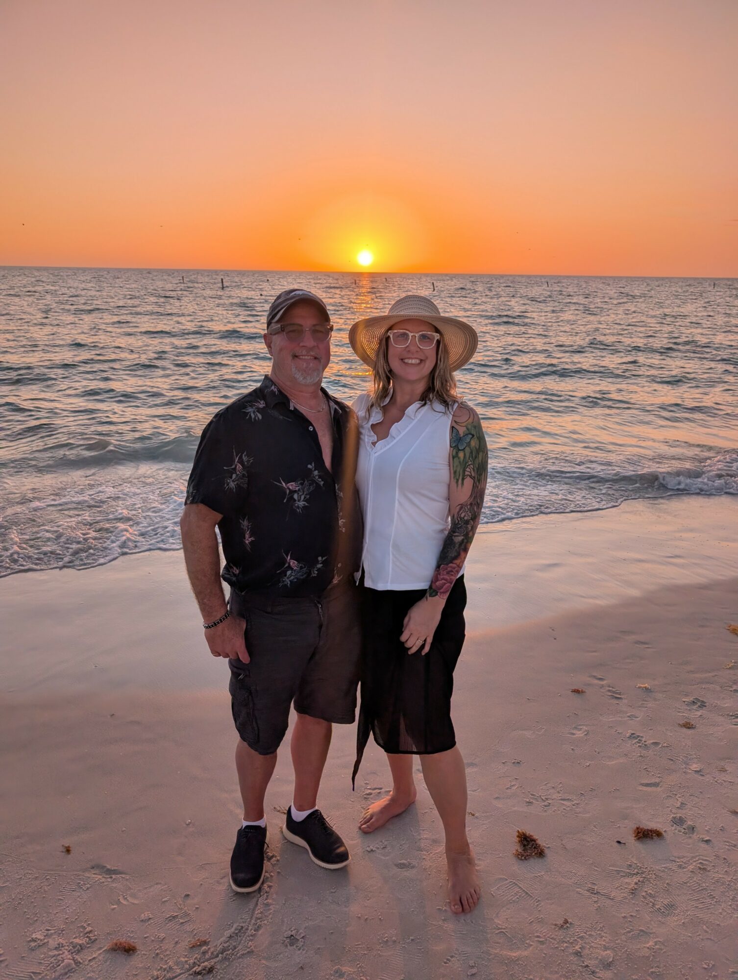 Two people stand on a beach at sunset, smiling, with the ocean and setting sun in the background.