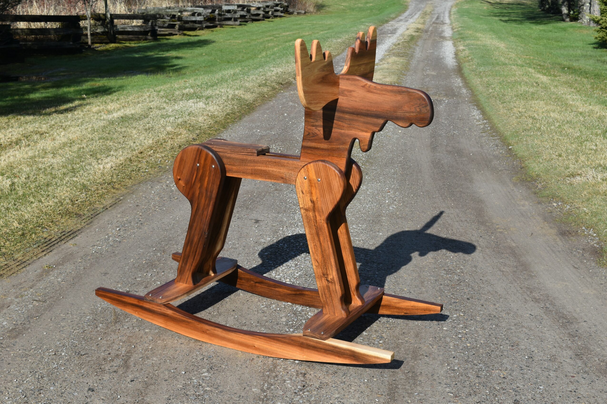 Wooden rocking horse on a gravel path with grass and trees in the background.