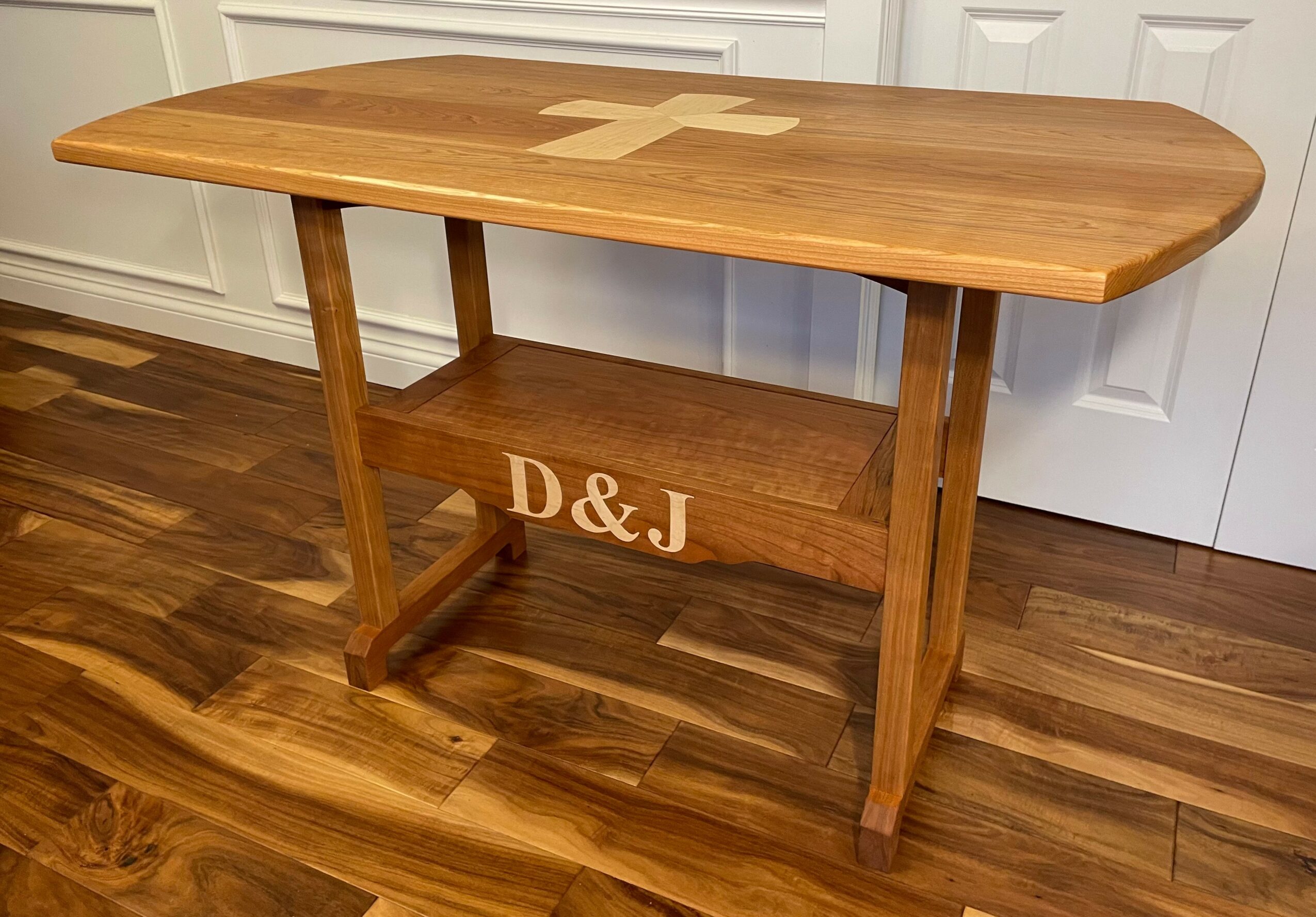 Wooden table with a cross-shaped inlay on top and a shelf with initials D&J underneath, on a hardwood floor.