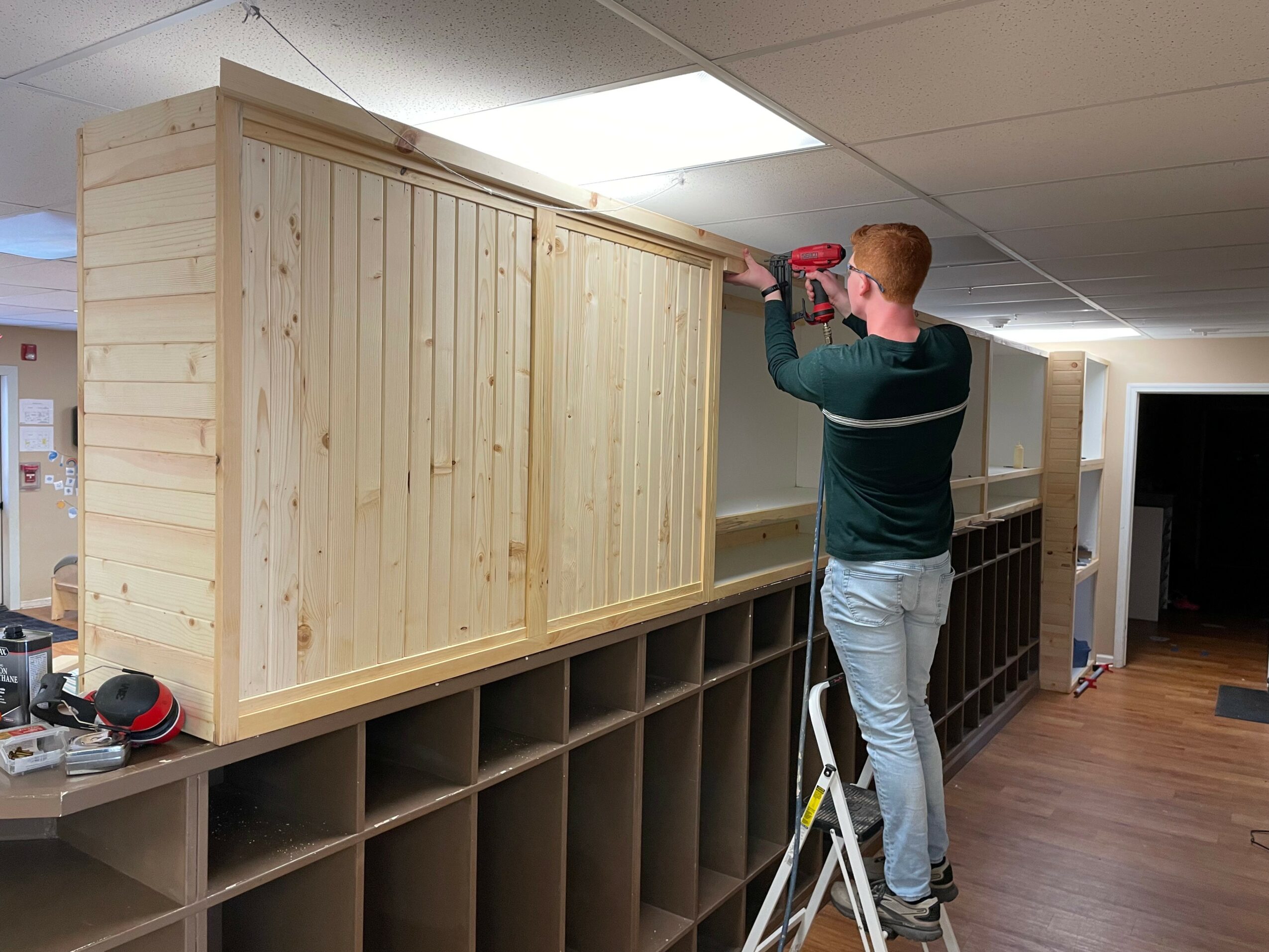 Person installing wooden panel on wall in indoor space, with storage cubbies below, ceiling lights above, and a ladder nearby.