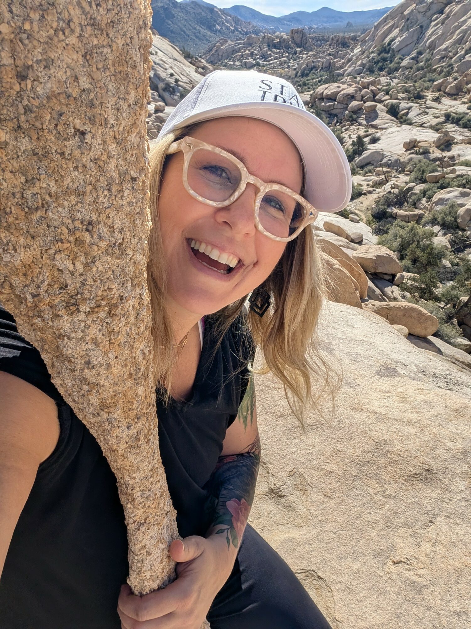 Person smiling, wearing glasses and a white cap, peeking from behind a large rock in a rocky desert landscape.