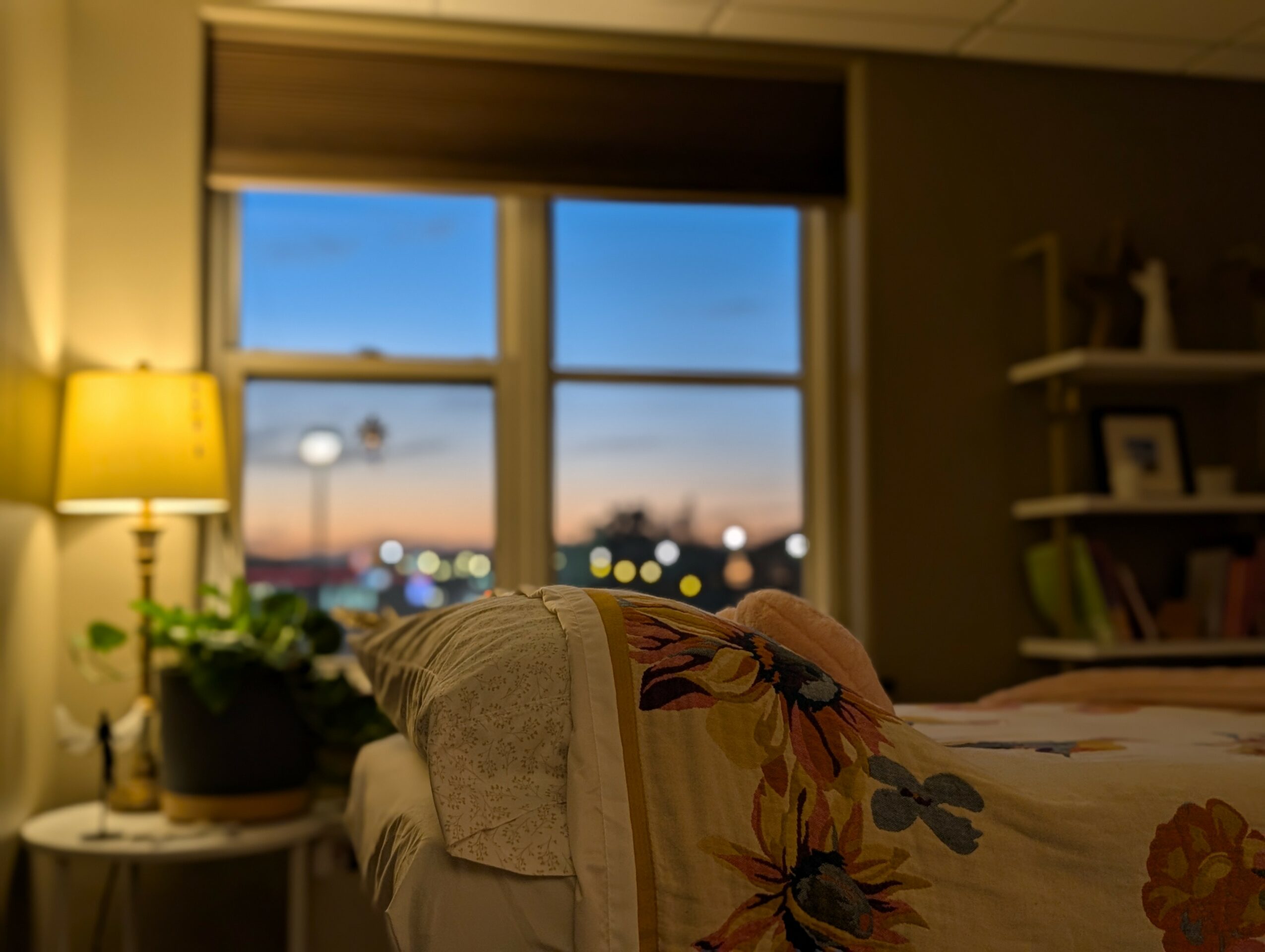 Bedroom with window showing a sunset sky, bed with floral bedding, lamp, and bookshelf, interior warmly lit.