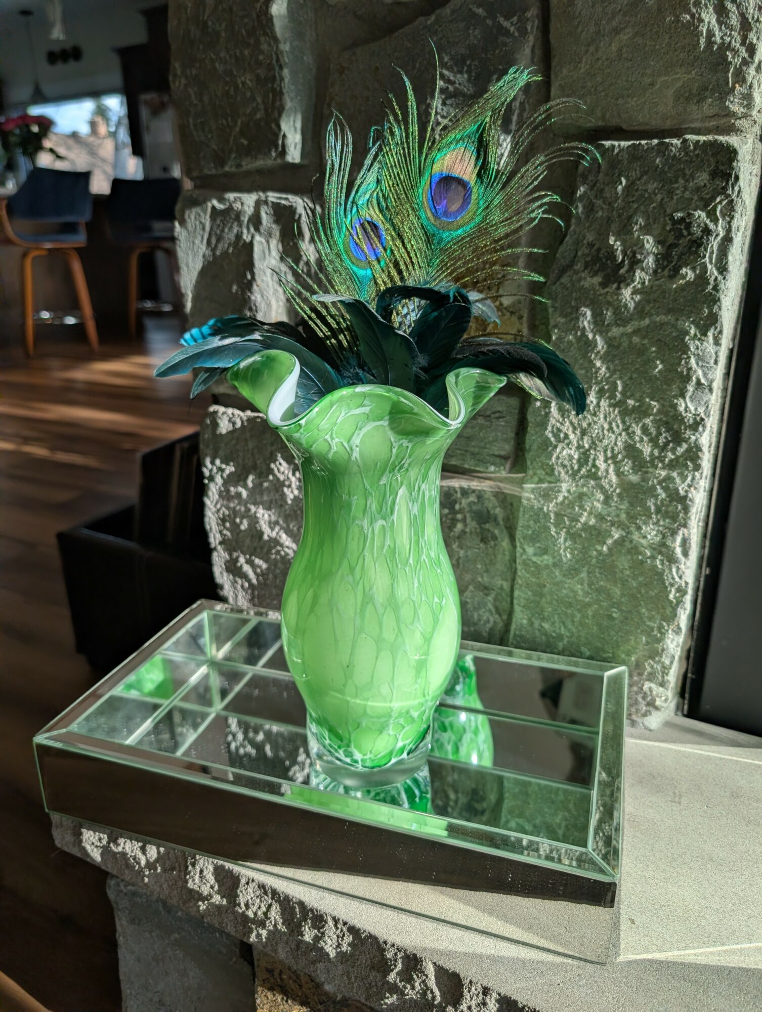 Green vase with peacock feathers on a mirrored tray against a stone wall, interior setting, natural light.