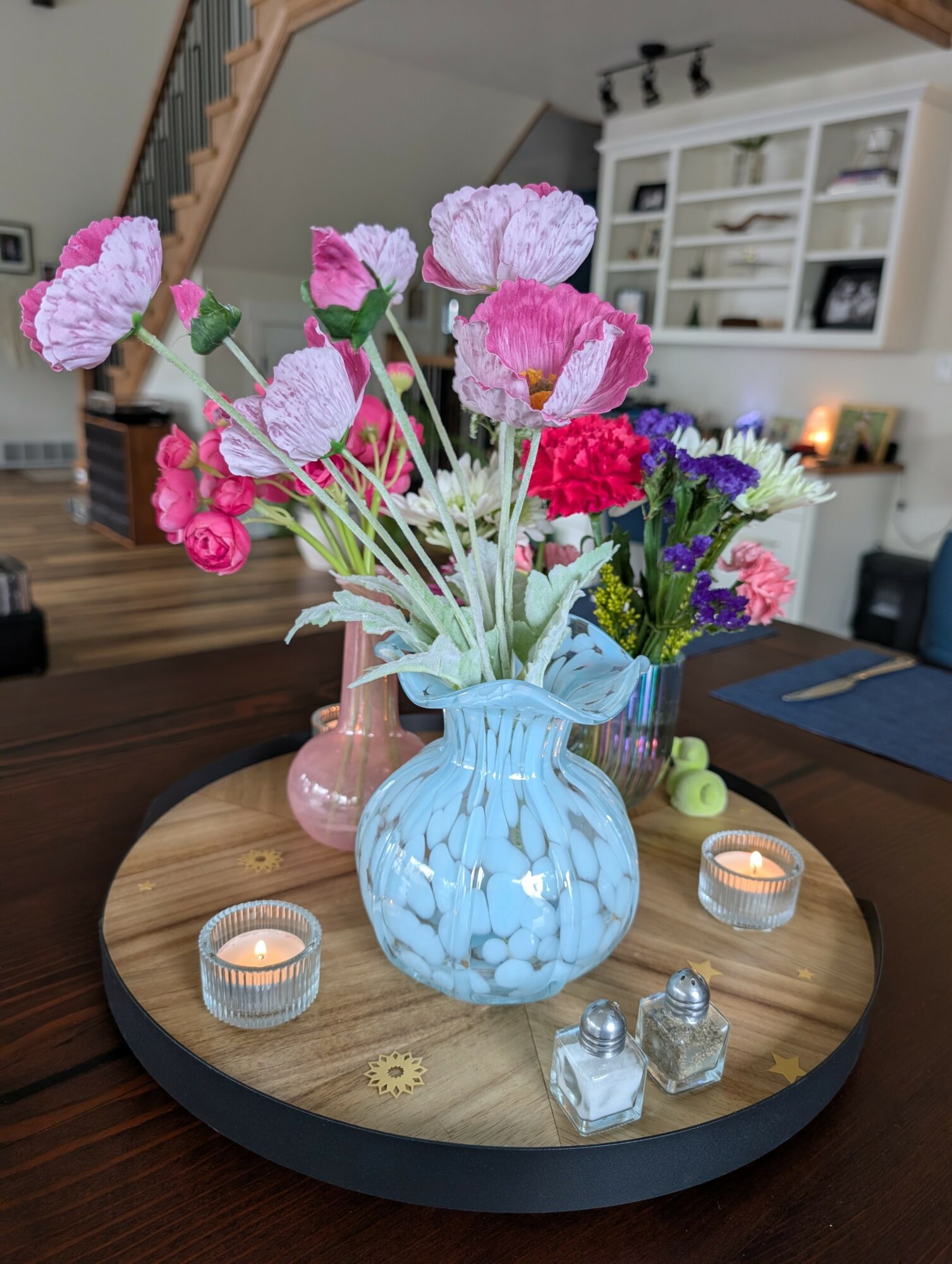 Colorful flowers in a blue vase on a round wooden tray with candles and decorative items, in a cozy living room.