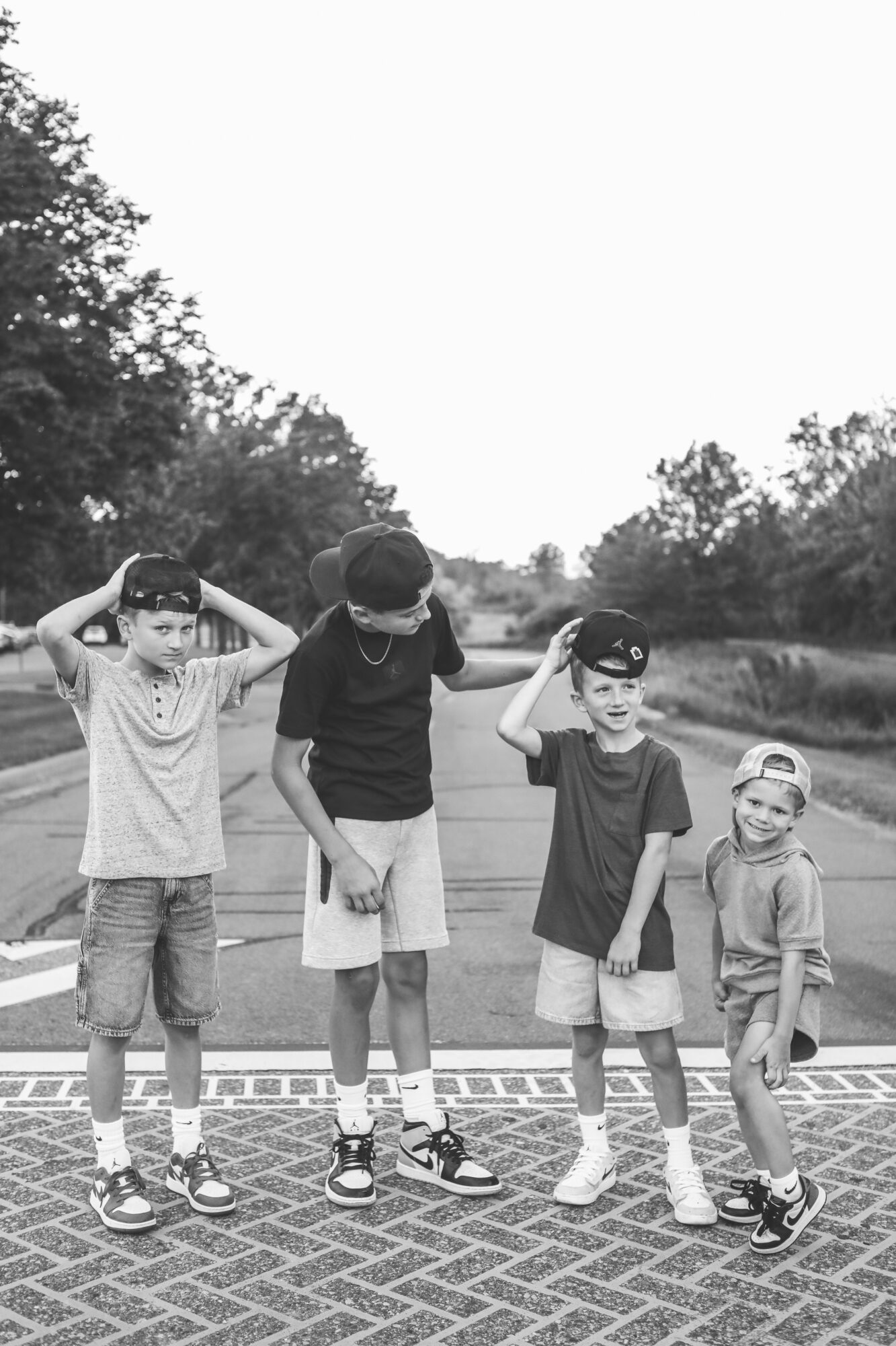 Four children standing outdoors on a paved surface, with trees and sky in the background, some saluting or adjusting their hats.