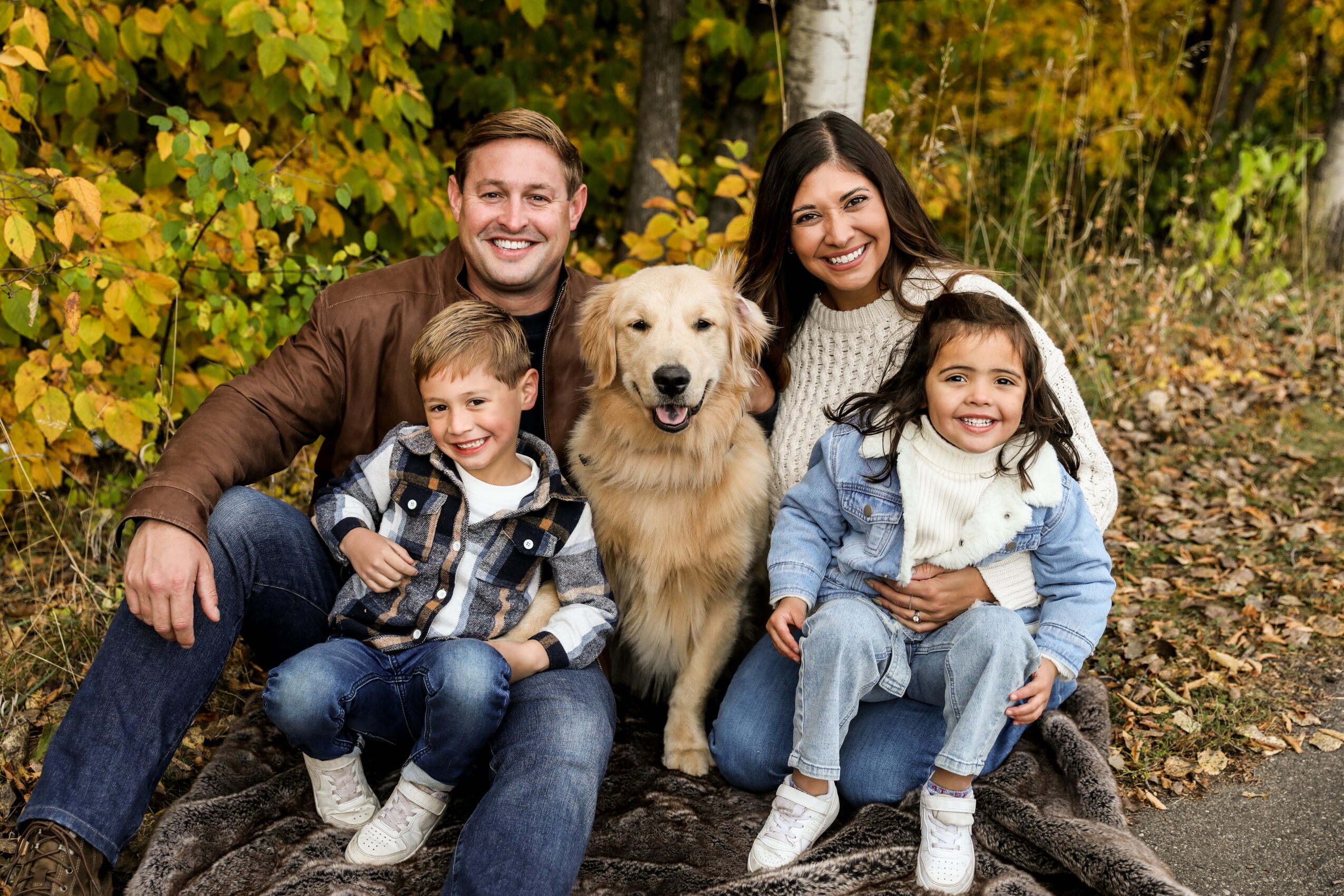 Family of four with a dog outdoors in autumn, sitting on a trail surrounded by colorful trees.