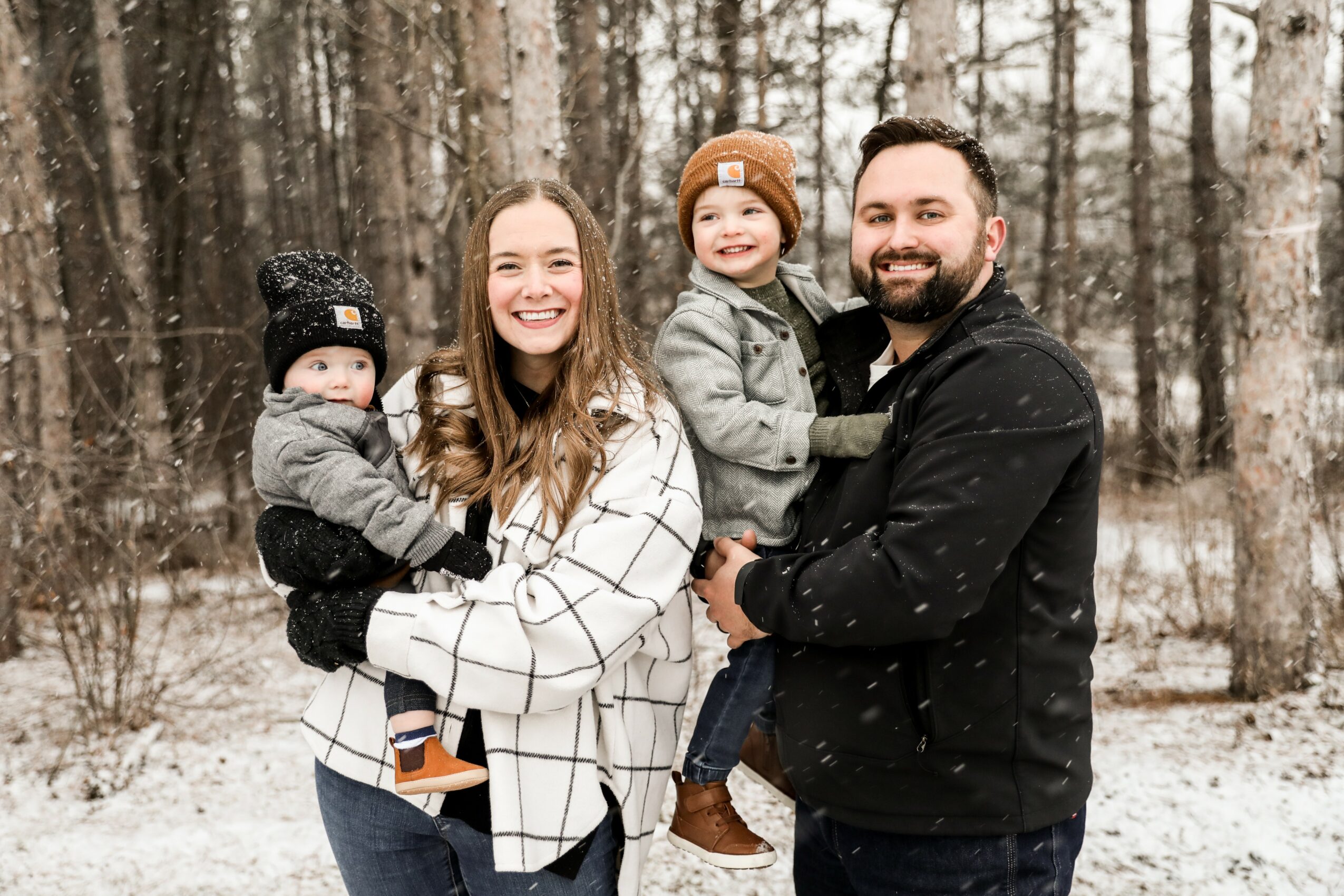 Family of four smiling outdoors in a snowy forest, two children held by adults, winter clothing, trees in background.