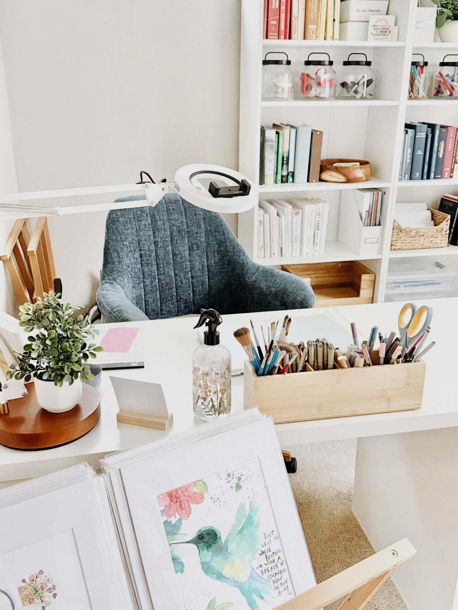 Office desk with a gray chair, potted plant, open book, and a wooden container with art supplies, against a white bookshelf.
