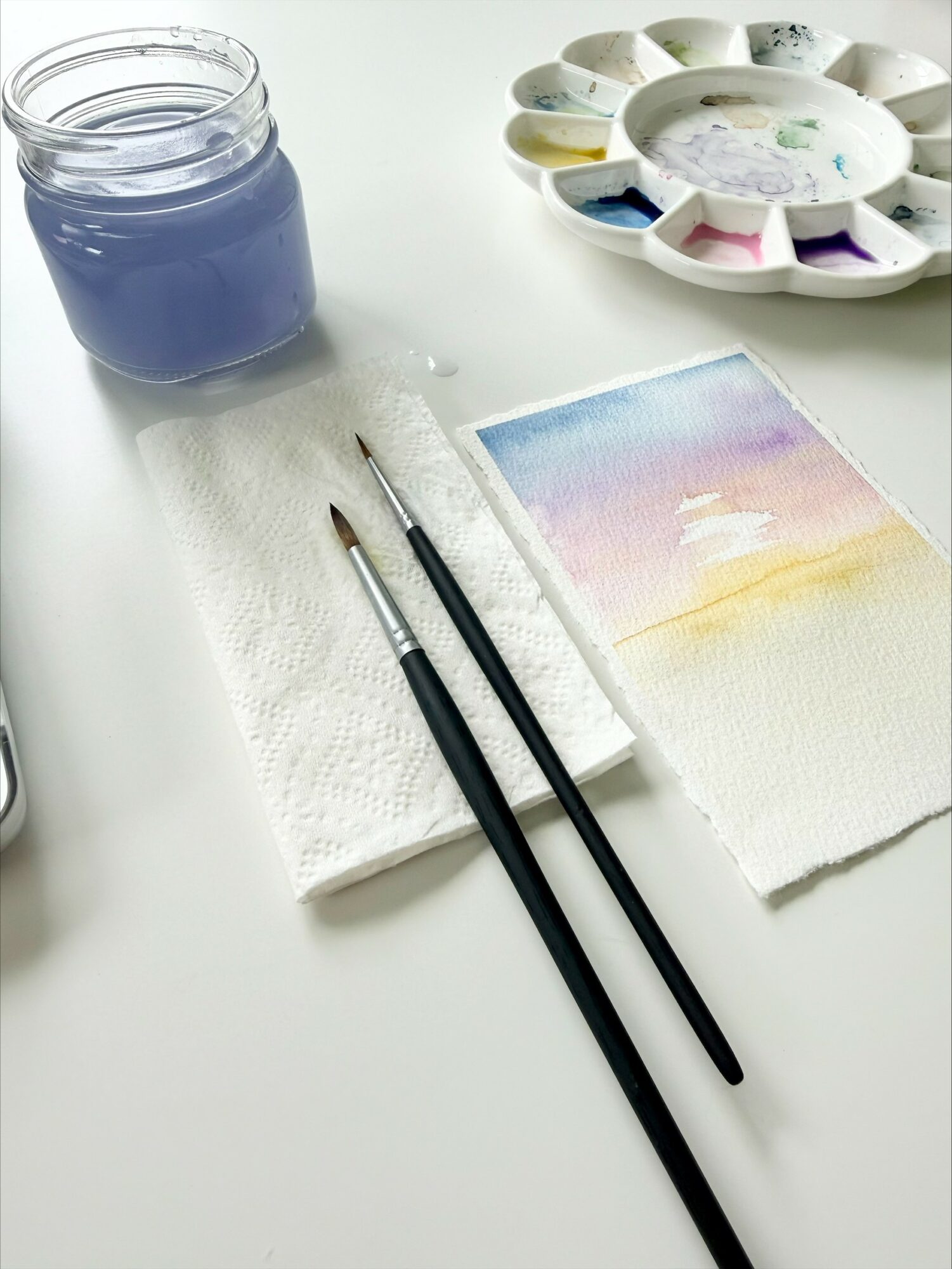 Watercolor painting supplies including brushes, a palette, a jar of blue paint, and a partially painted paper on a white surface.