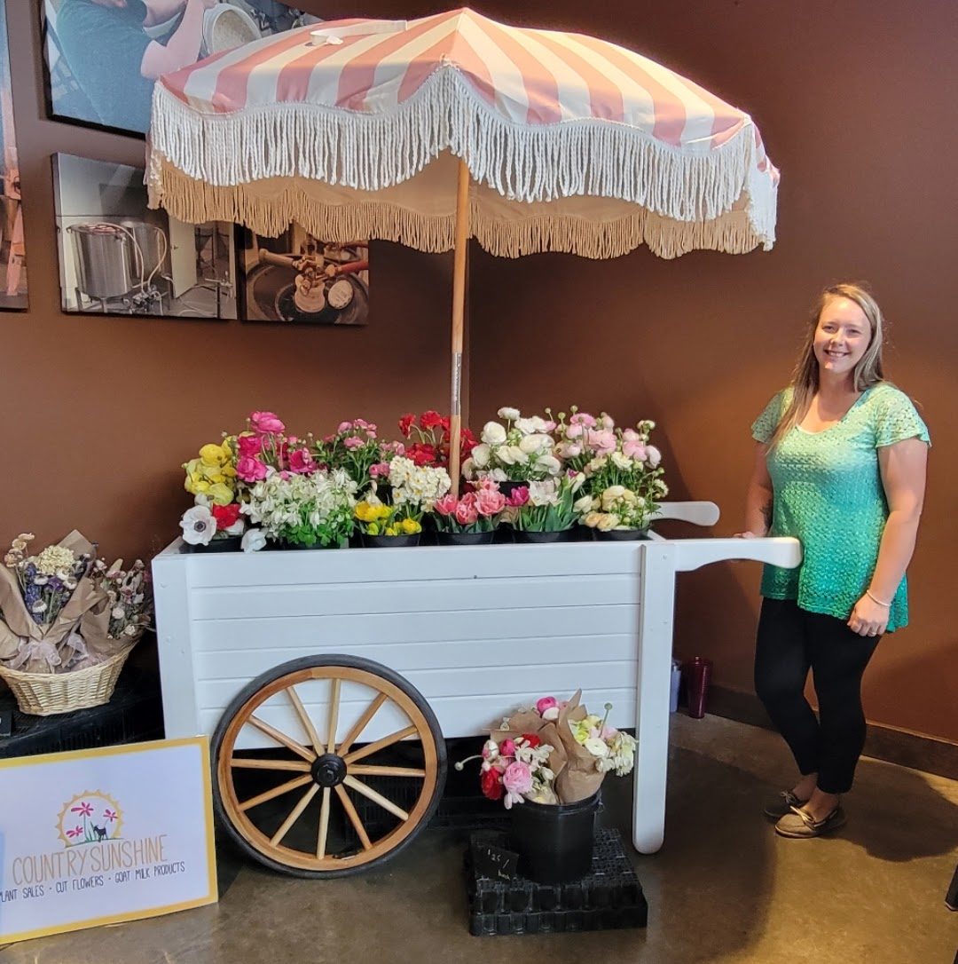 Woman standing next to a flower cart with an umbrella, flowers, and a sign, indoors. She is smiling and wearing a green top.