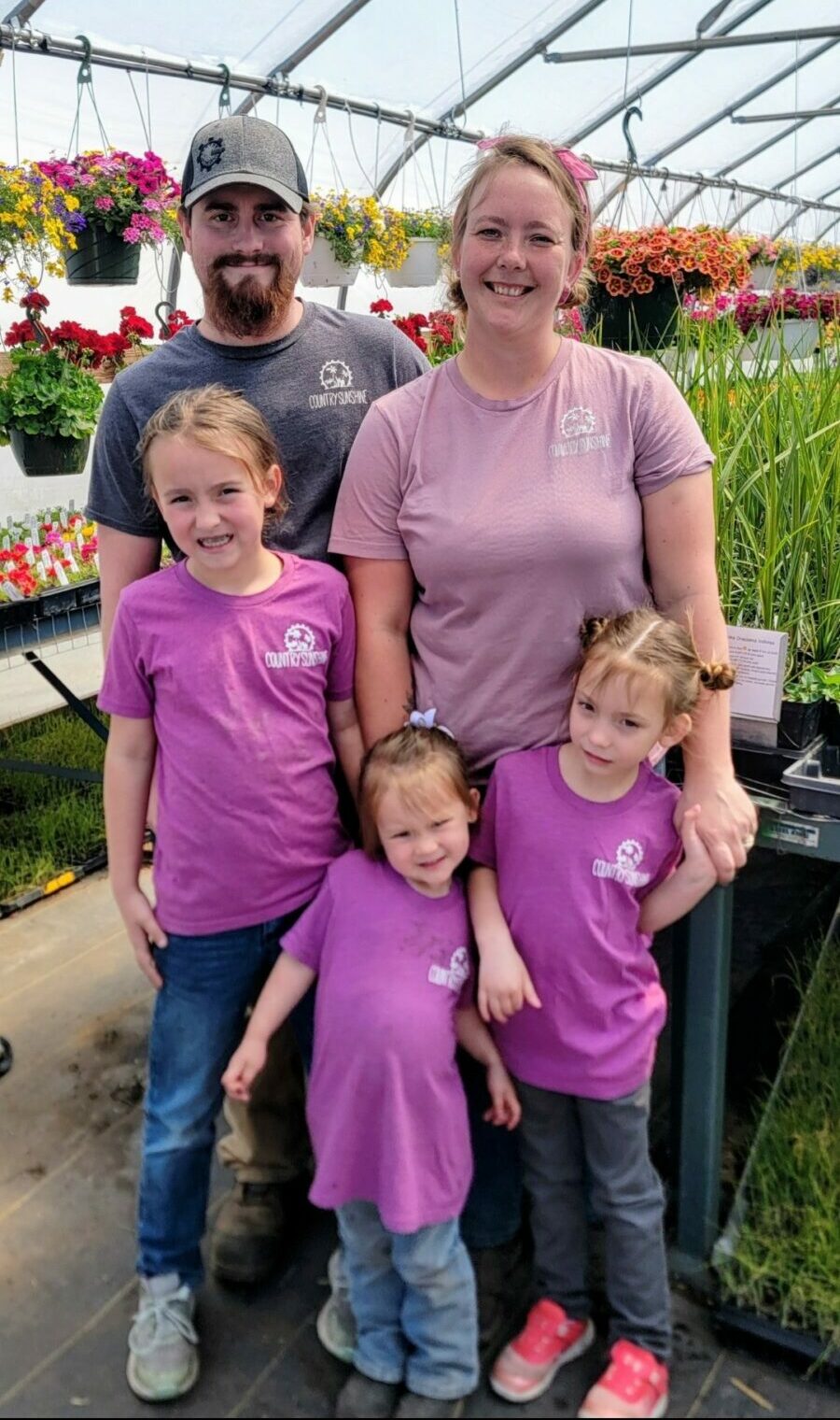 Family of five standing in a greenhouse with hanging flowers, smiling, with children in purple shirts and an adult woman in pink