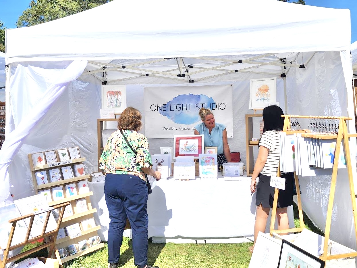 Two women browse art prints at a booth with a man behind the table, under a white canopy, outdoor setting.