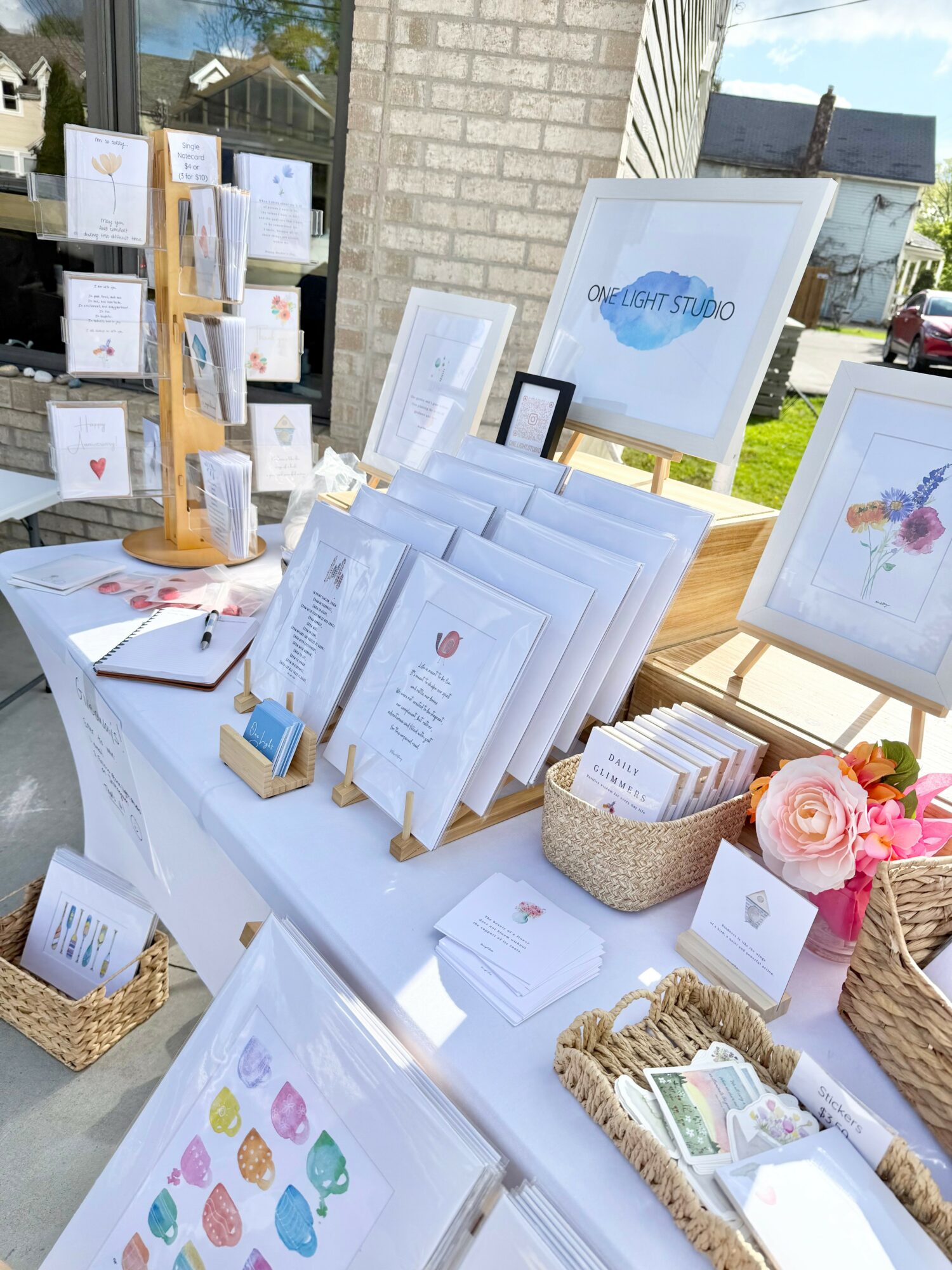 Outdoor market stall displays framed artwork, printed materials, and decorative items on a white table with baskets and flowers.