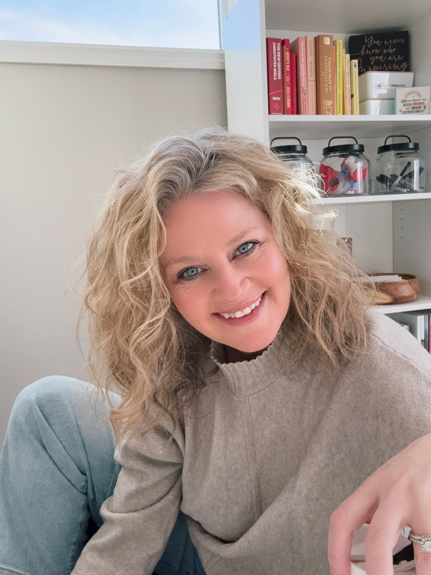 Smiling woman with curly blonde hair sitting indoors near a bookshelf, wearing a beige sweater.