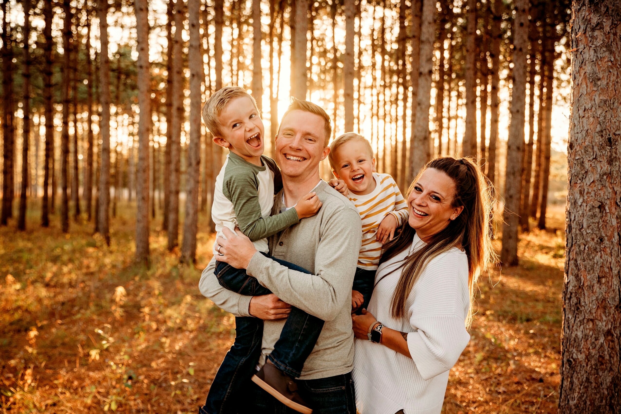 Family of four smiling in a forest during sunset, with trees and sunlight in the background.
