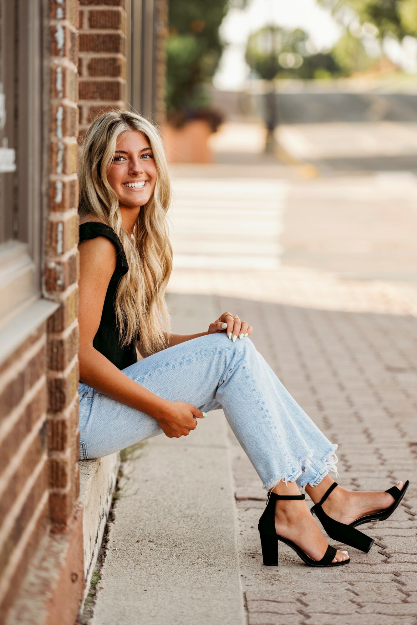Young woman with long blonde hair sitting on a sidewalk, smiling, wearing black top, light jeans, and black heels.