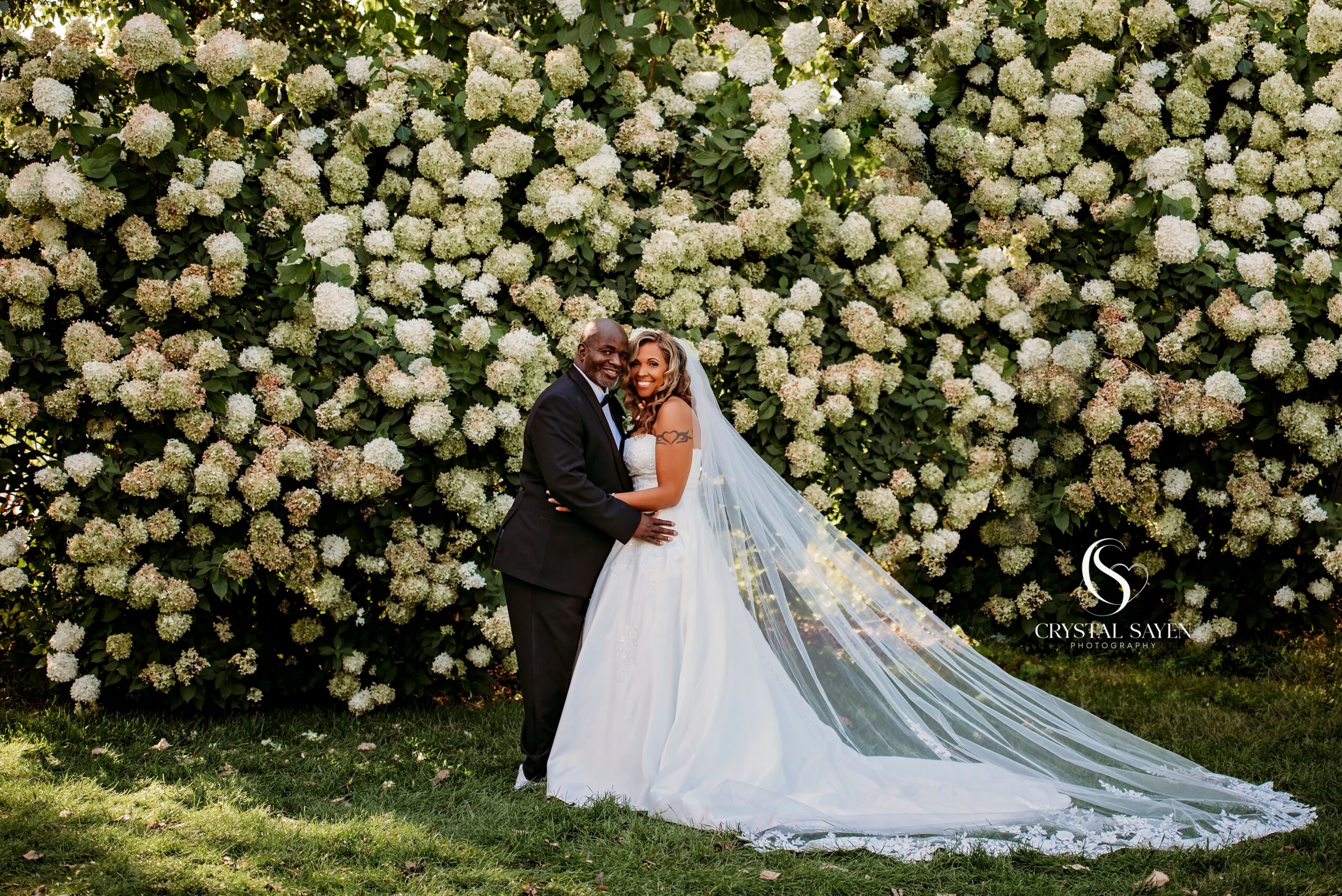 A bride and groom stand together outdoors in front of a large wall of white flowers, smiling and holding hands.