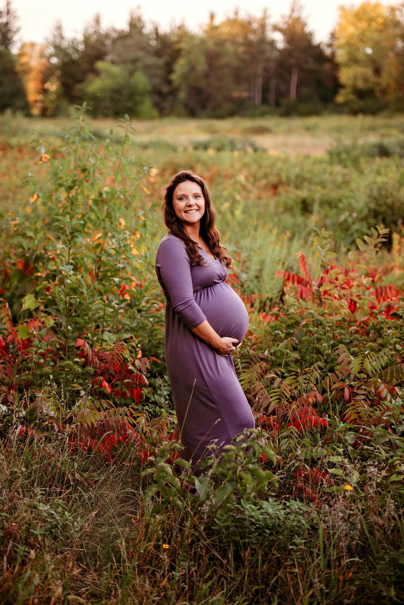 Pregnant woman standing in a field with colorful autumn foliage, smiling, wearing a purple dress.
