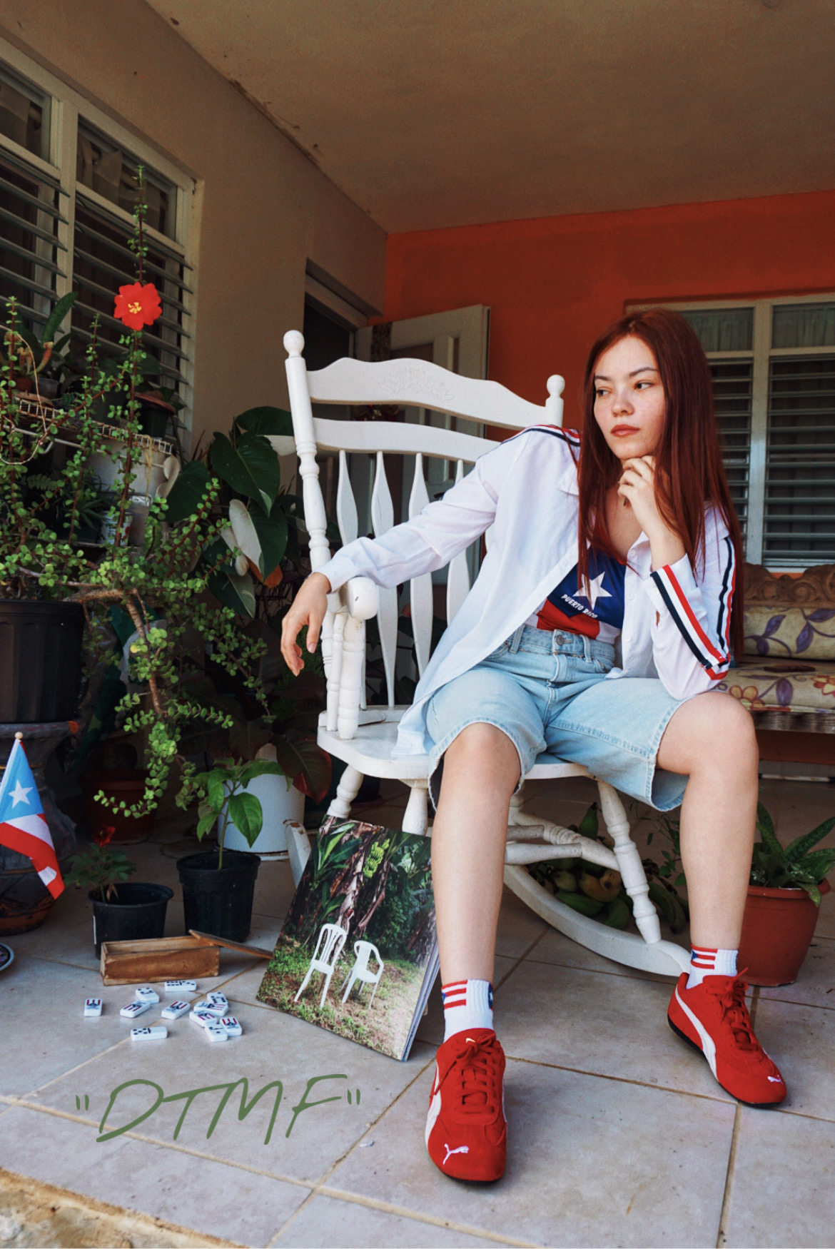 Young woman with long hair sitting on a white rocking chair in a room with plants and windows, wearing a white jacket, denim shorts, and red sneakers.