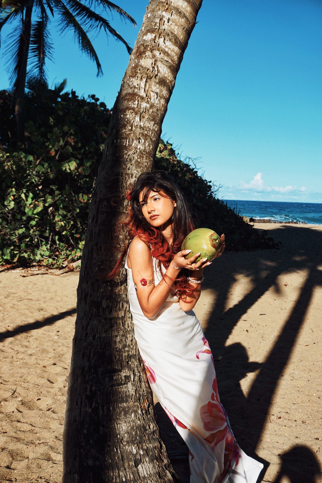 Young woman in white dress holding a coconut on a beach with palm trees and ocean in background.