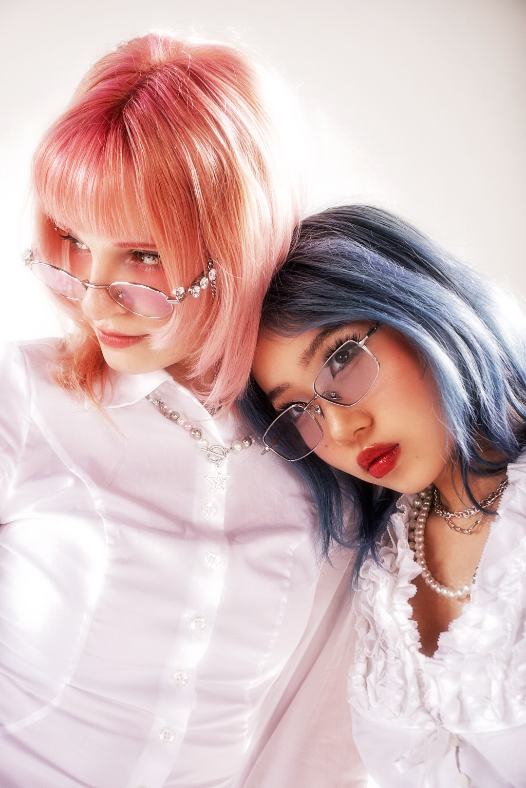 Two women with colorful hair and glasses, one with pink hair and the other with blue hair, pose closely together.