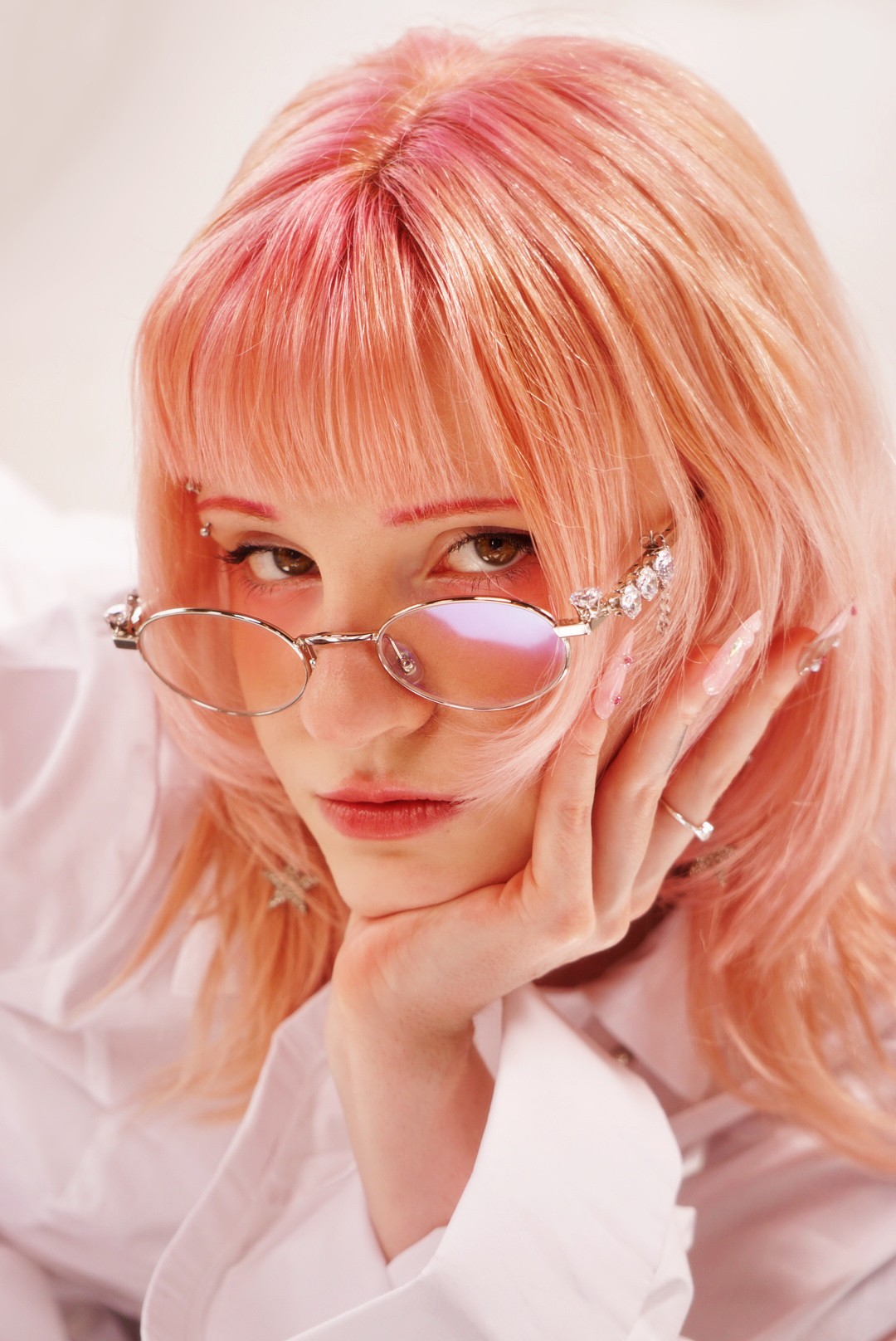 Person with pink hair and glasses resting chin on hand, looking at camera.