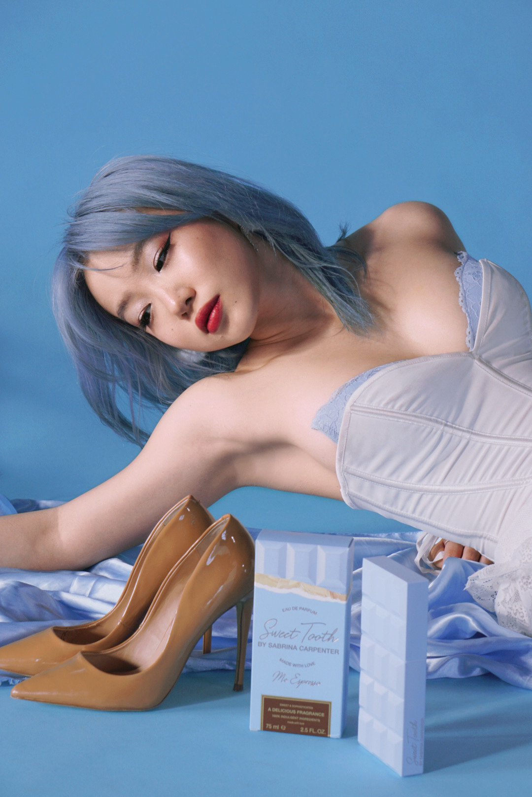 Woman with blue hair lying on her side, wearing a strapless top, with brown high heels and gift boxes nearby.
