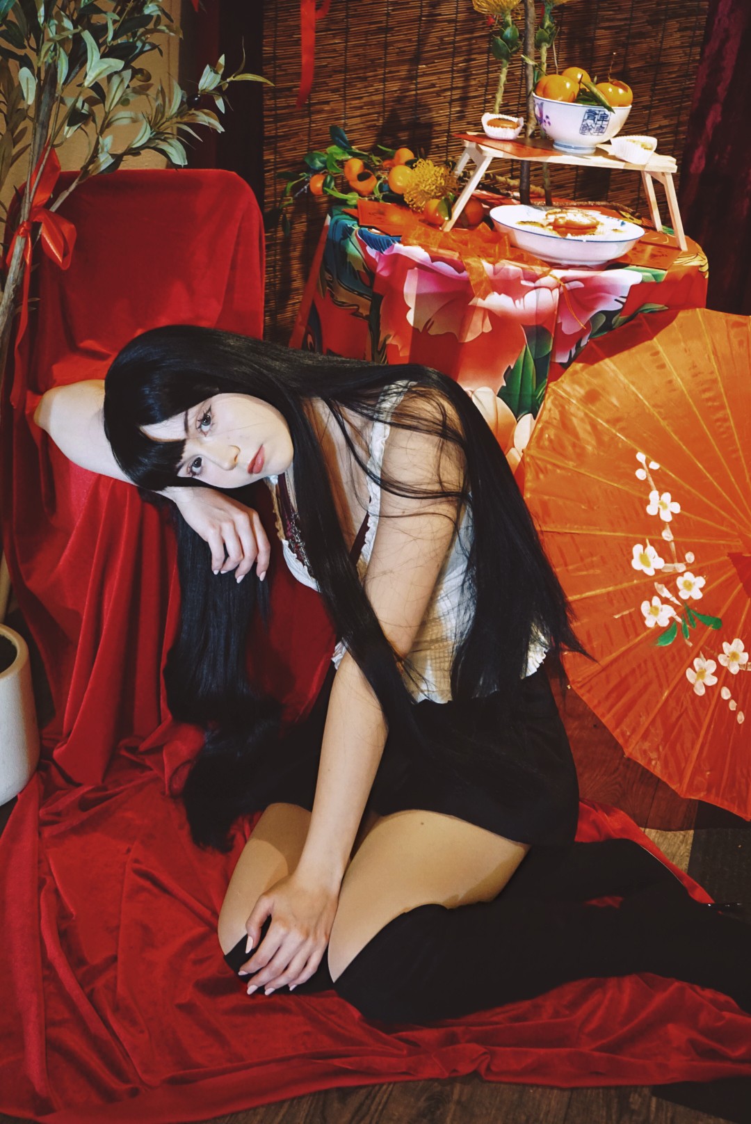 Young woman with long black hair, sitting on red fabric, resting her head on her hand, surrounded by Asian-themed decor.