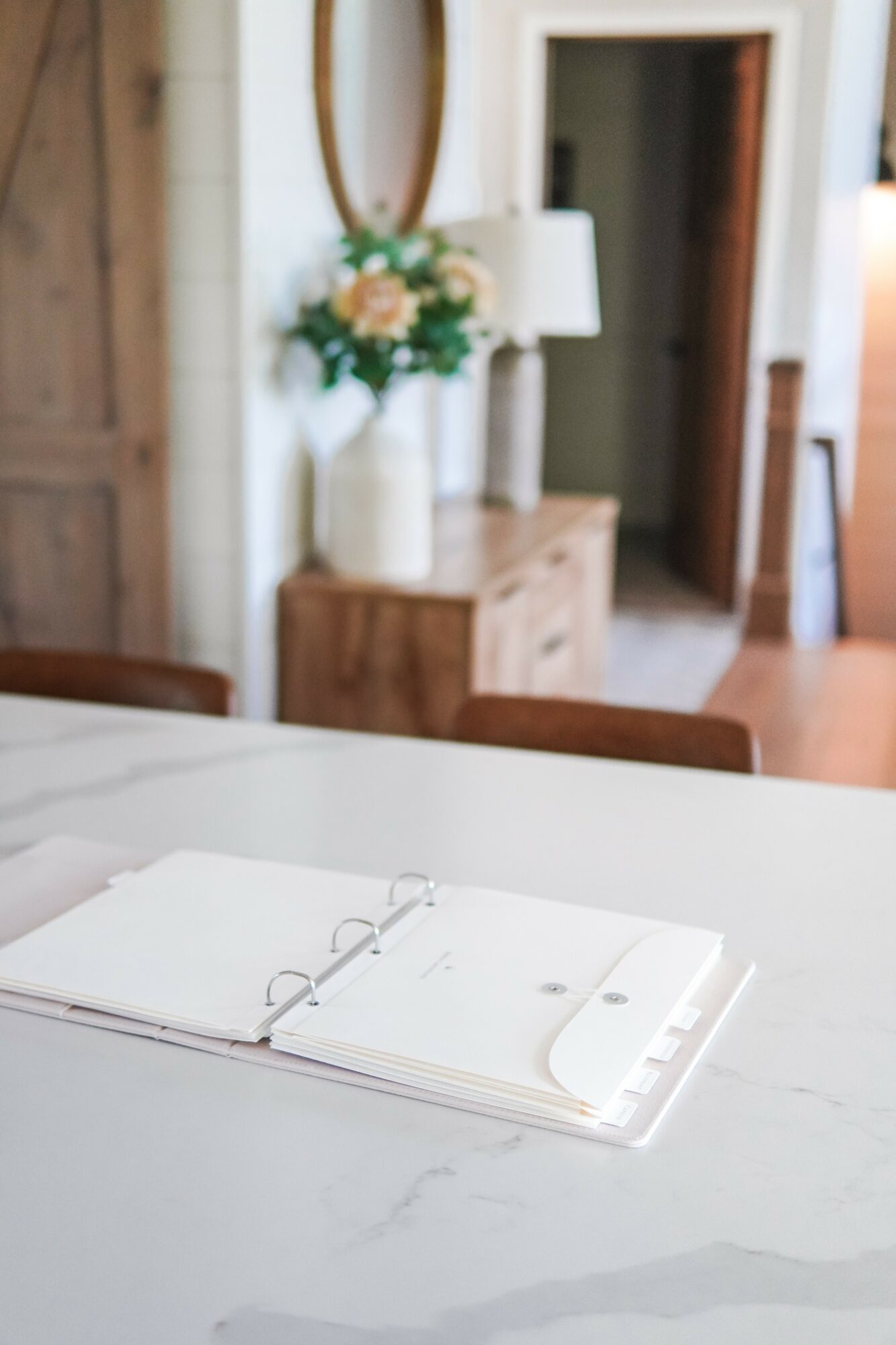 Open notebook on a white table with a blurred background of a bedroom, dresser, mirror, and flowers.