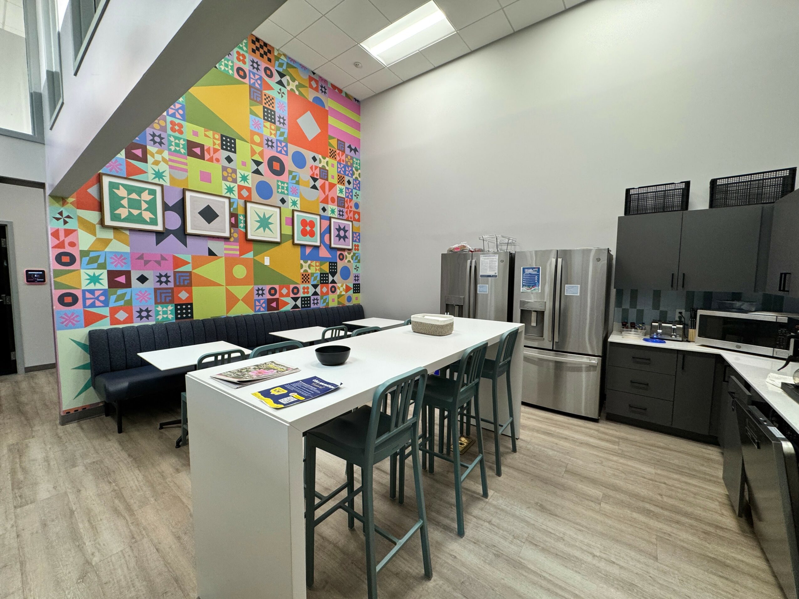 Meeting room with colorful geometric wall, white table, black chairs, and kitchen appliances in the background.
