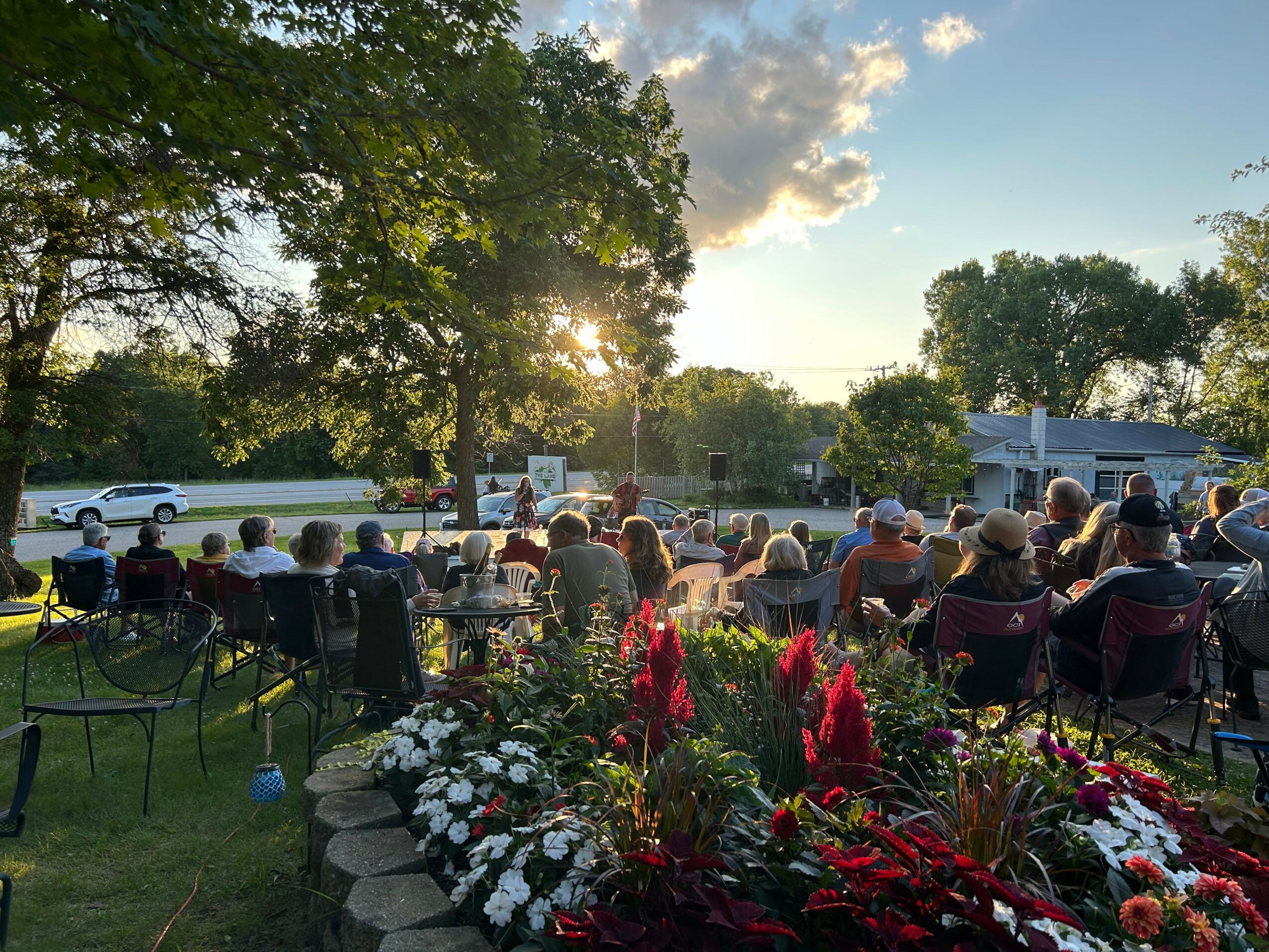 Group of people gathered outdoors in a park-like setting with trees and flowers, watching a sunset.