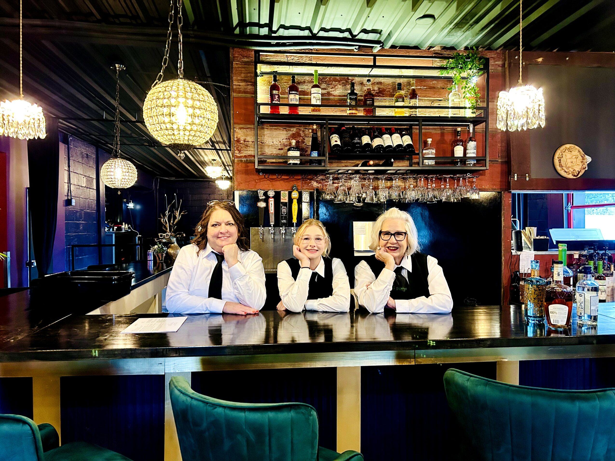 Three women sit at a bar counter in a dimly lit, modern bar with shelves of bottles behind them.